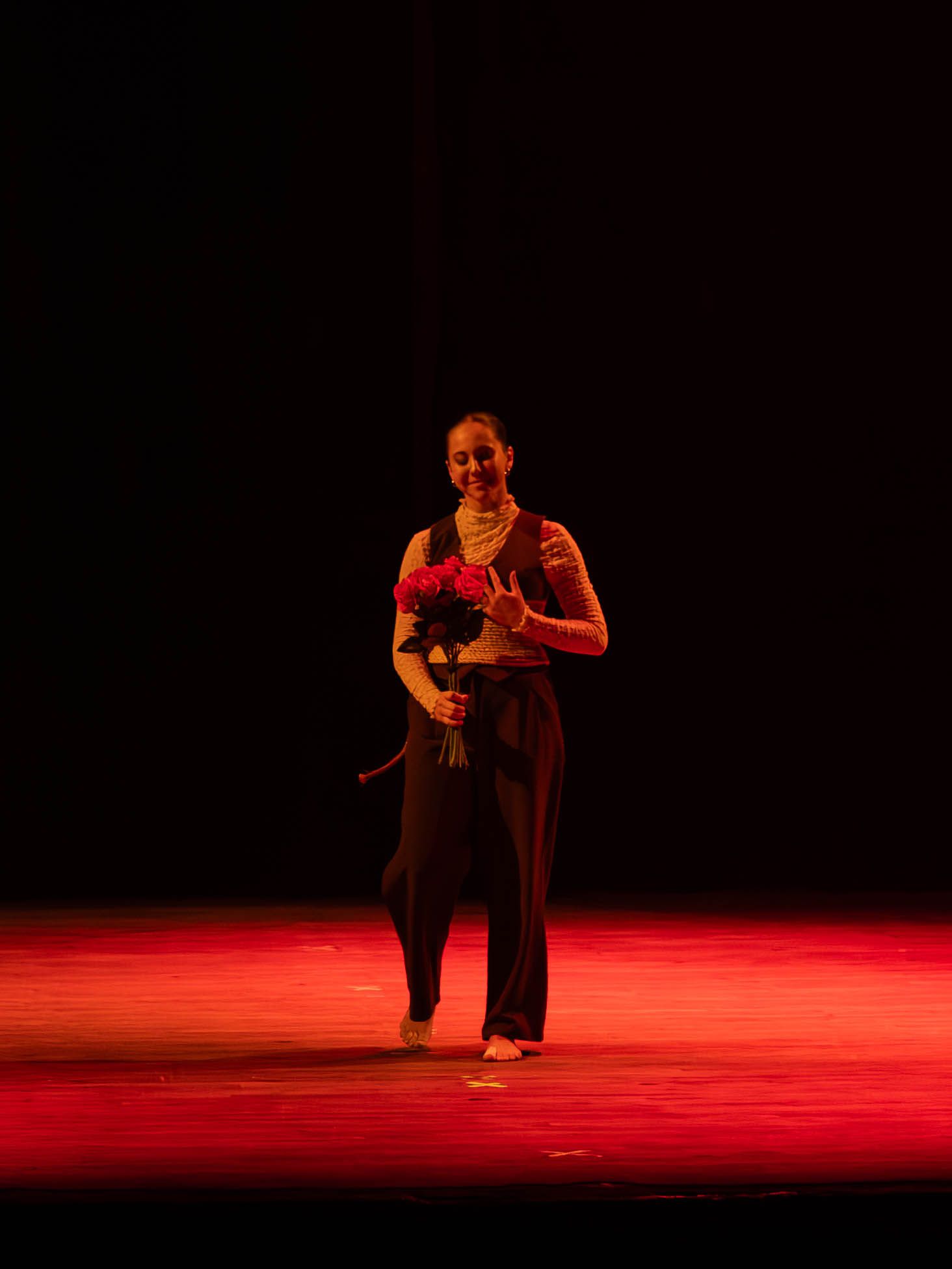 A dancer in black attire holds red roses on a dimly lit stage under red lighting after a performance.