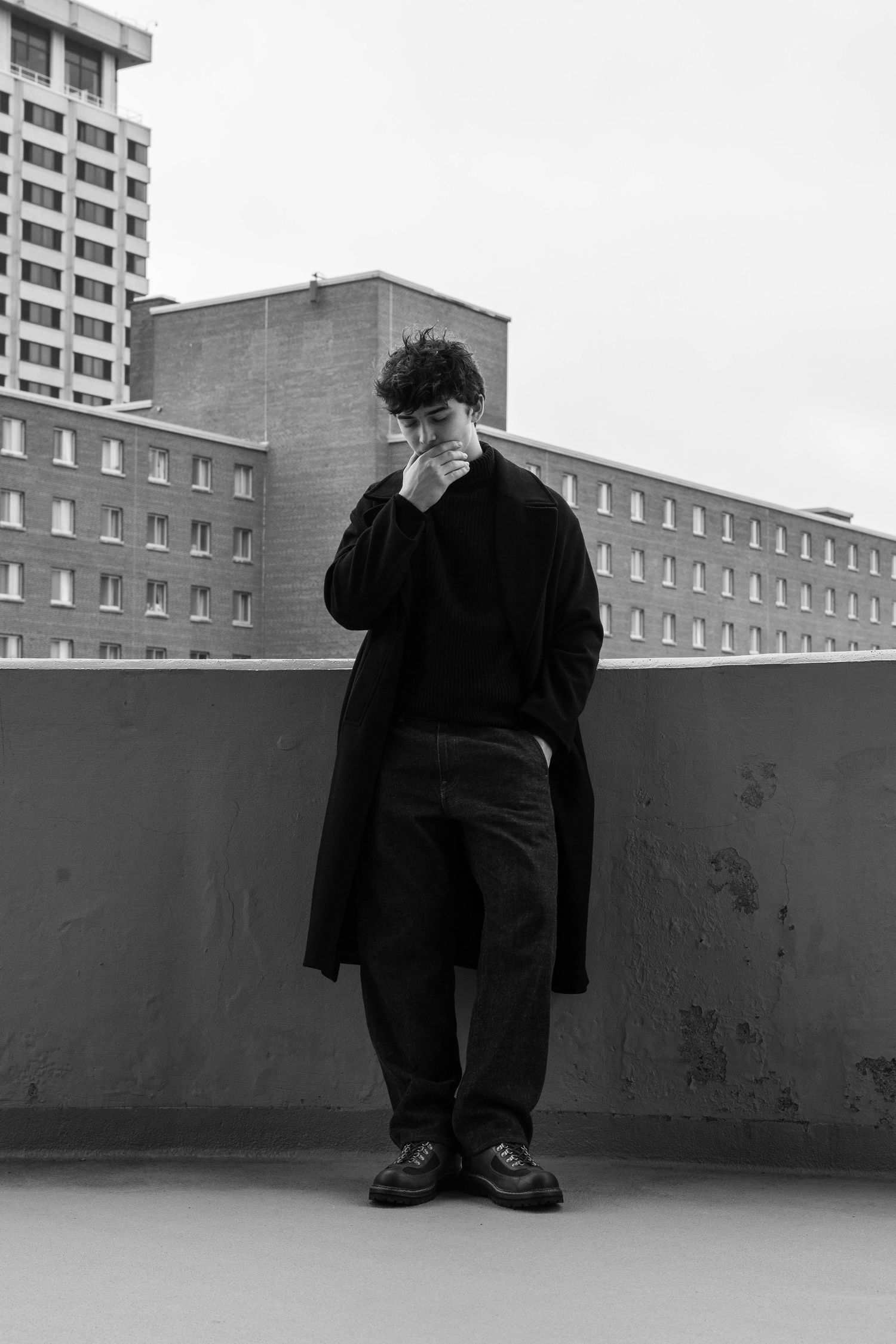 Black and white photo of a young man in a long dark coat standing on a concrete rooftop with brick buildings behind him.