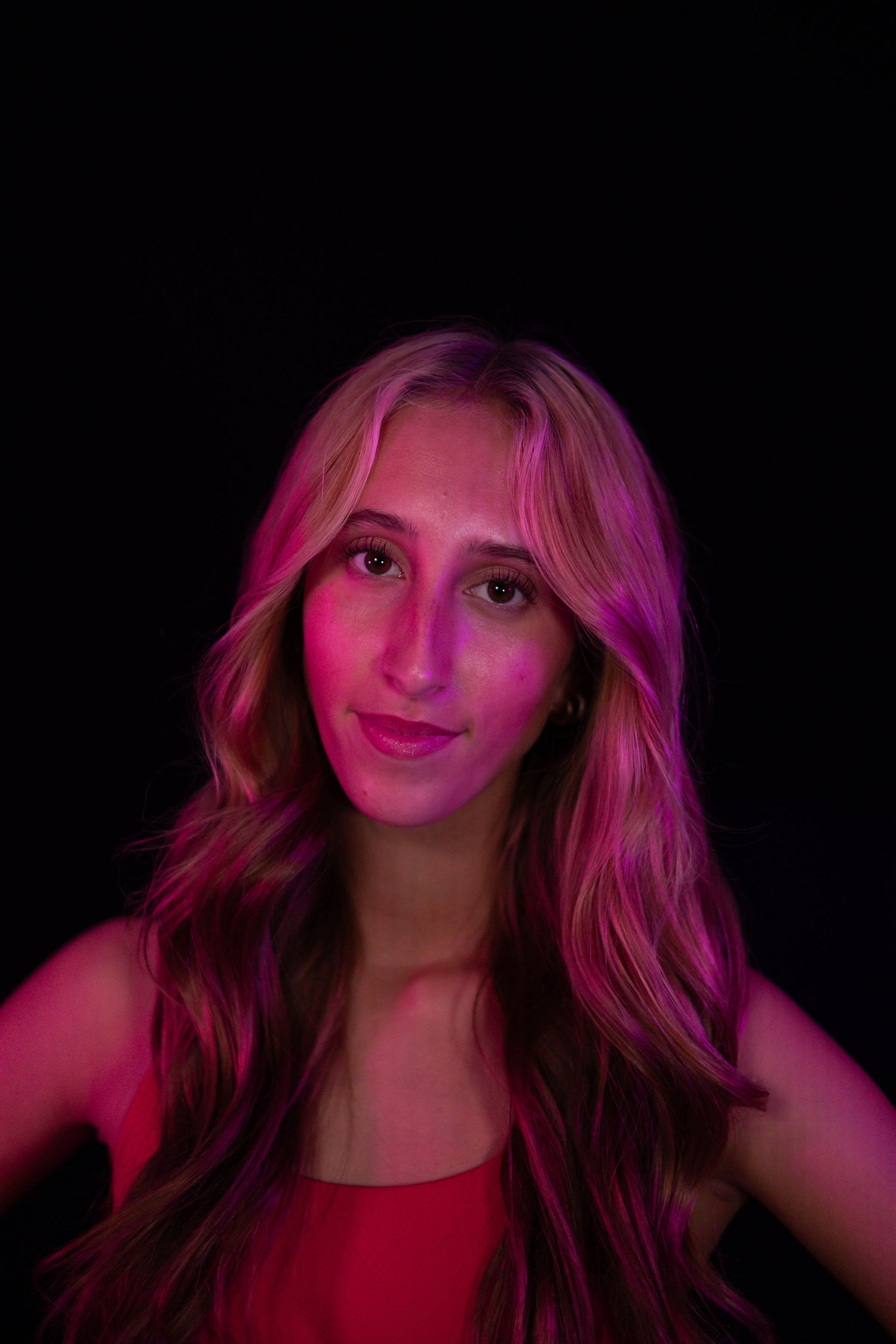 Young woman with long wavy blonde hair and a red top poses against a black background, lit by vivid pink and magenta studio lighting.