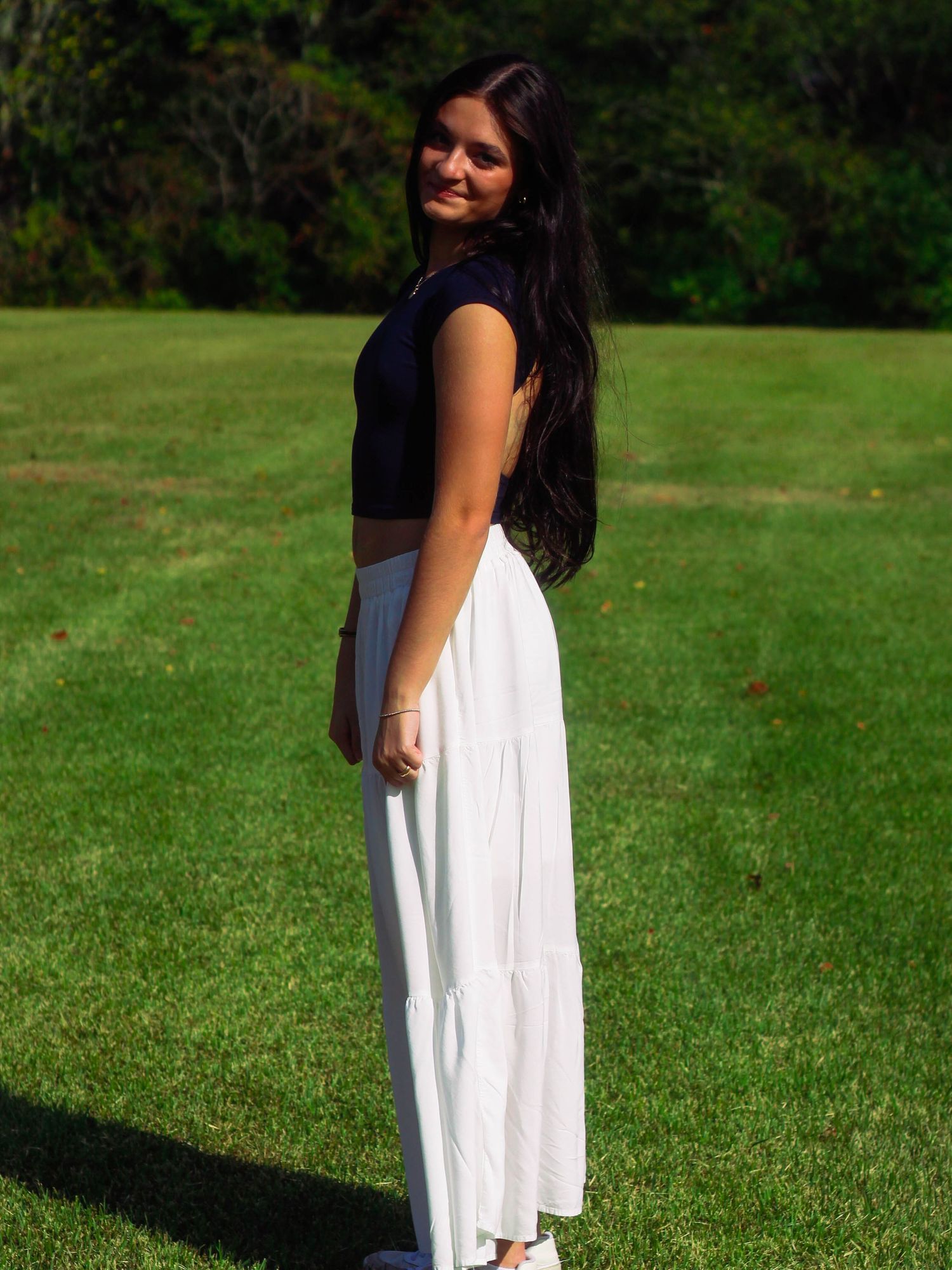 Young woman in black crop top and white tiered maxi skirt smiling over shoulder on green lawn.