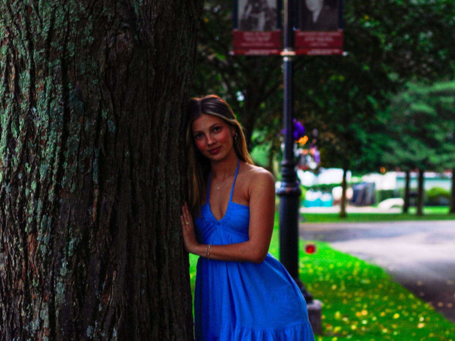 Young woman in blue spaghetti strap dress leaning against large tree trunk in park.