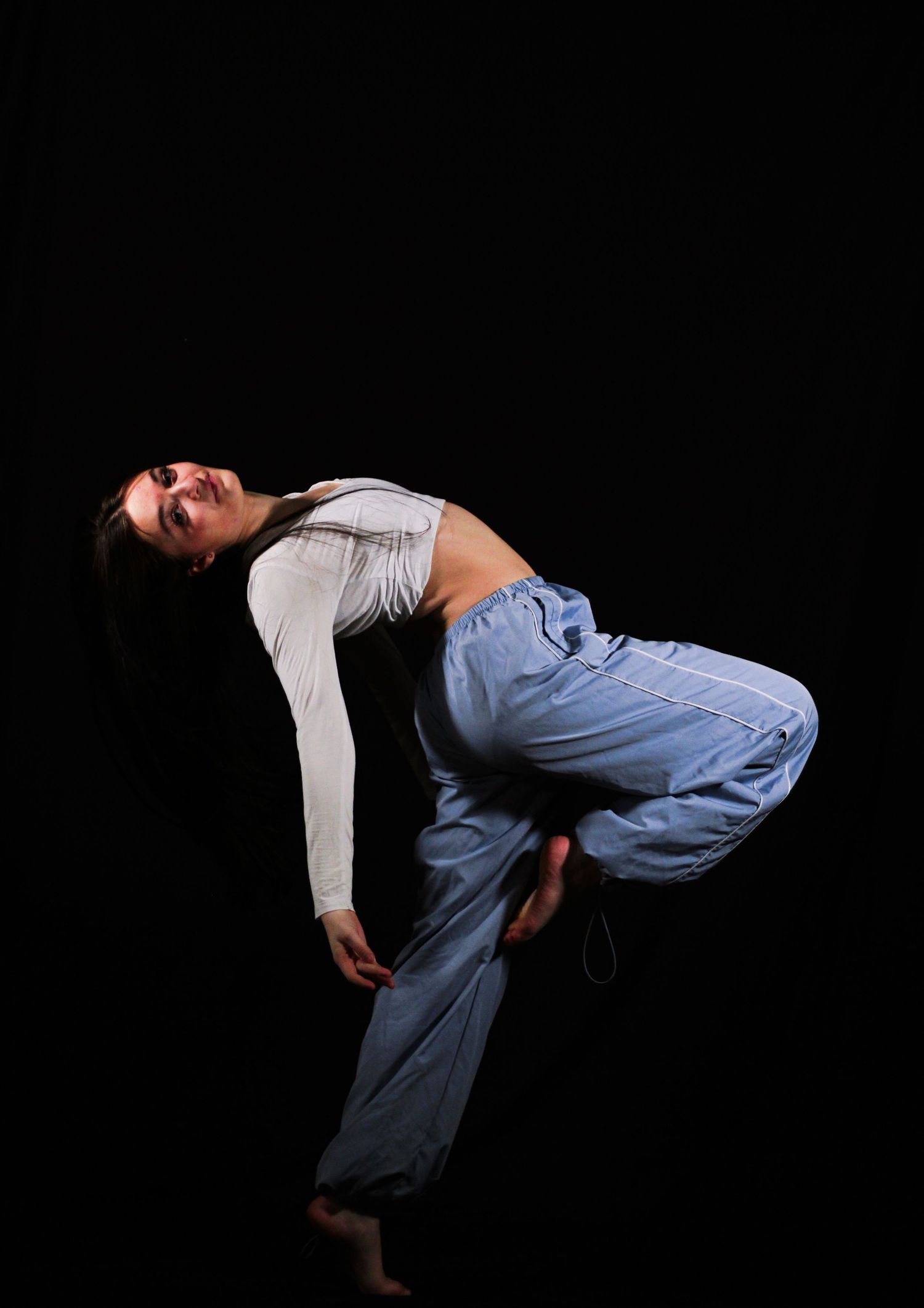 Barefoot dancer in light blue pants and a gray long-sleeve top arches backward in a dynamic pose against a black studio background.
