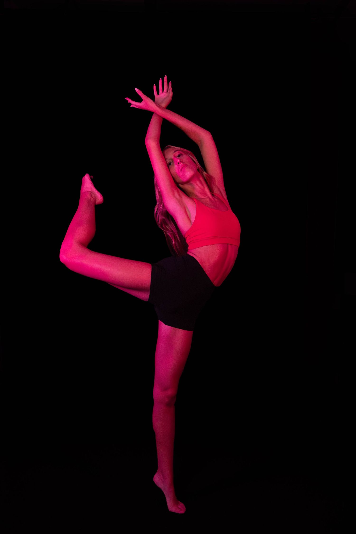 A dancer in a red top and black shorts balances on one leg in a high kick, arms raised, lit by vivid pink light against a black background.