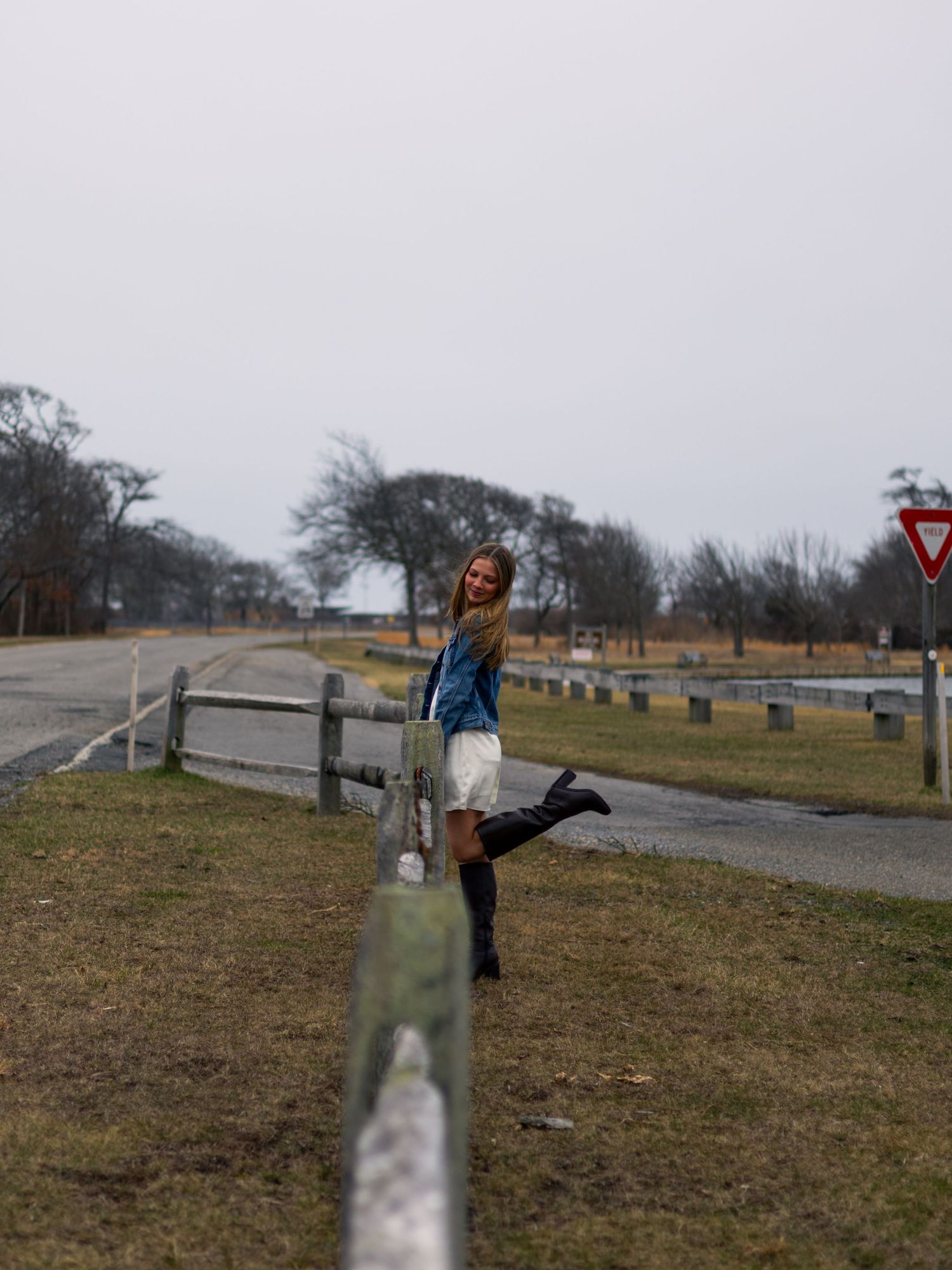 Smiling woman in a denim jacket, white skirt and black boots kicks one leg up beside a wooden fence near a road on an overcast day.