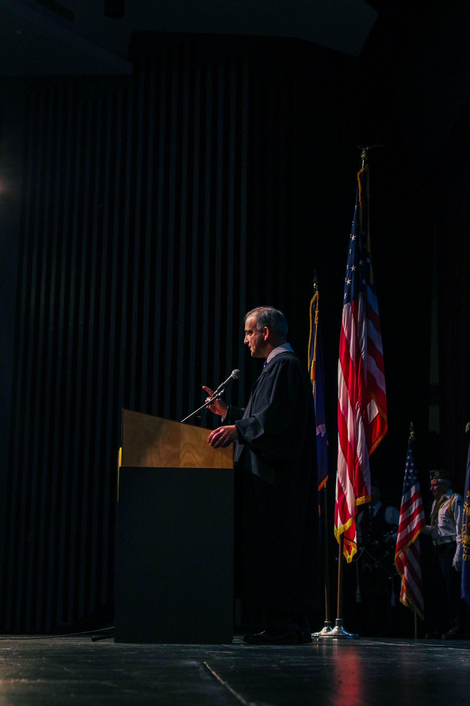 Speaker at podium on dark stage with American and military flags during a formal ceremony or graduation event.