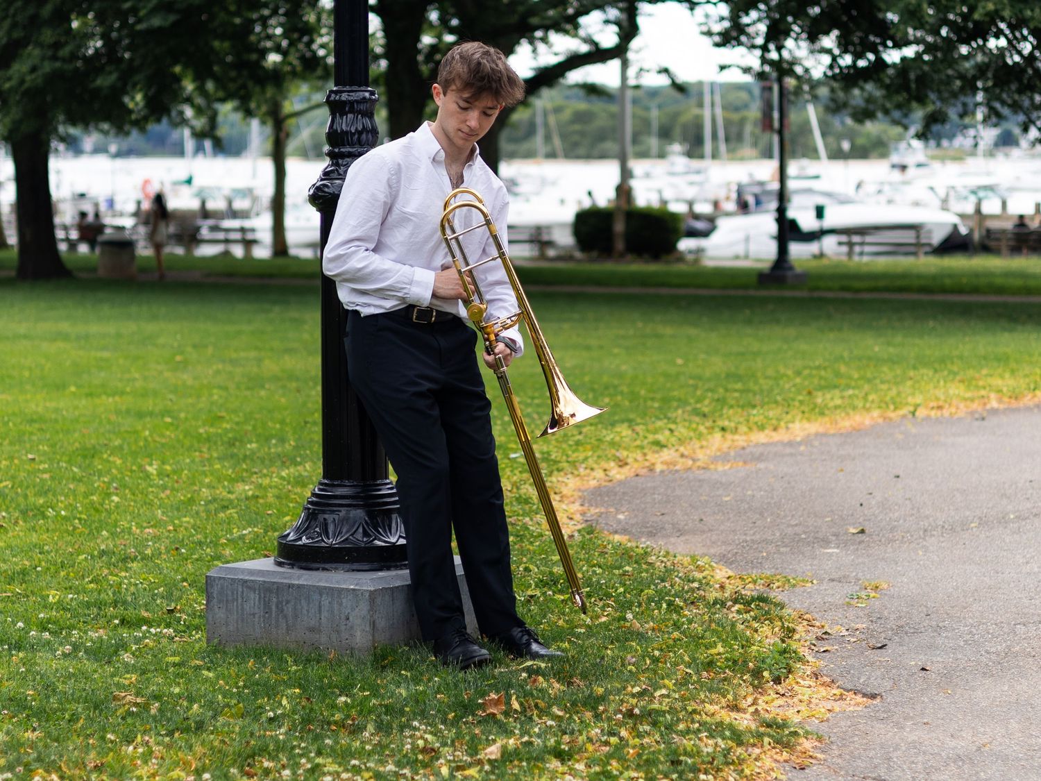 Young male musician in white shirt leaning on pedestal with trombone in park.