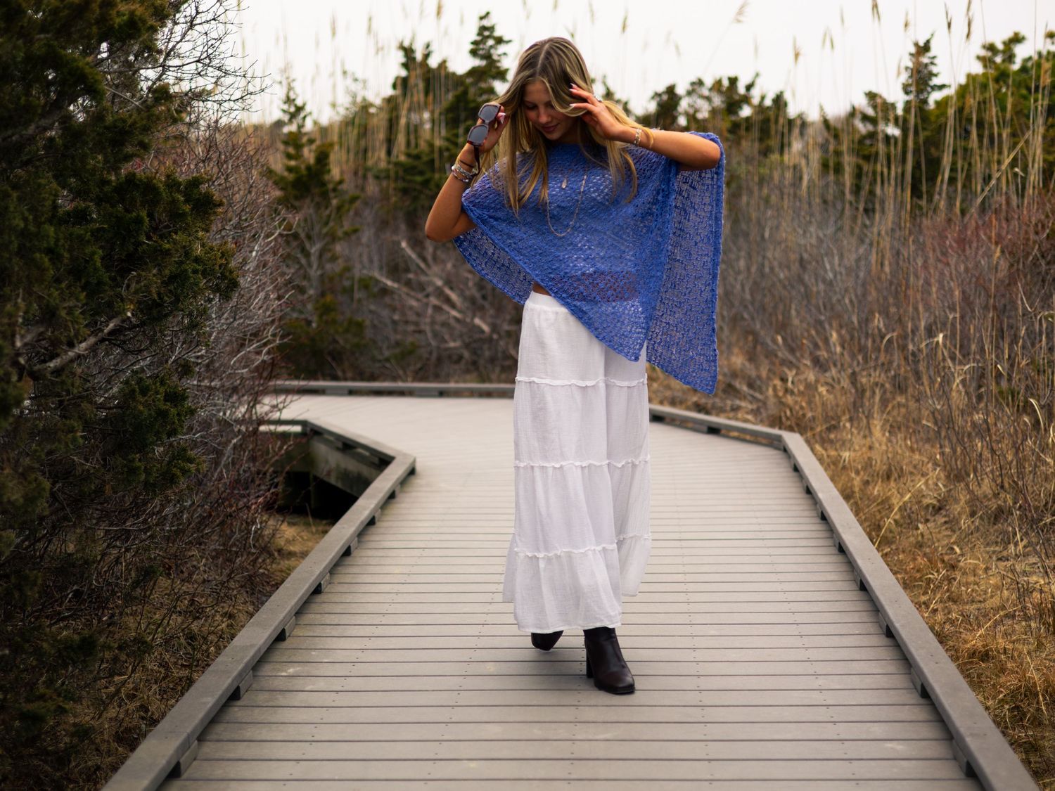 Woman in blue lace poncho and white maxi skirt walking on a wooden boardwalk through tall grass.