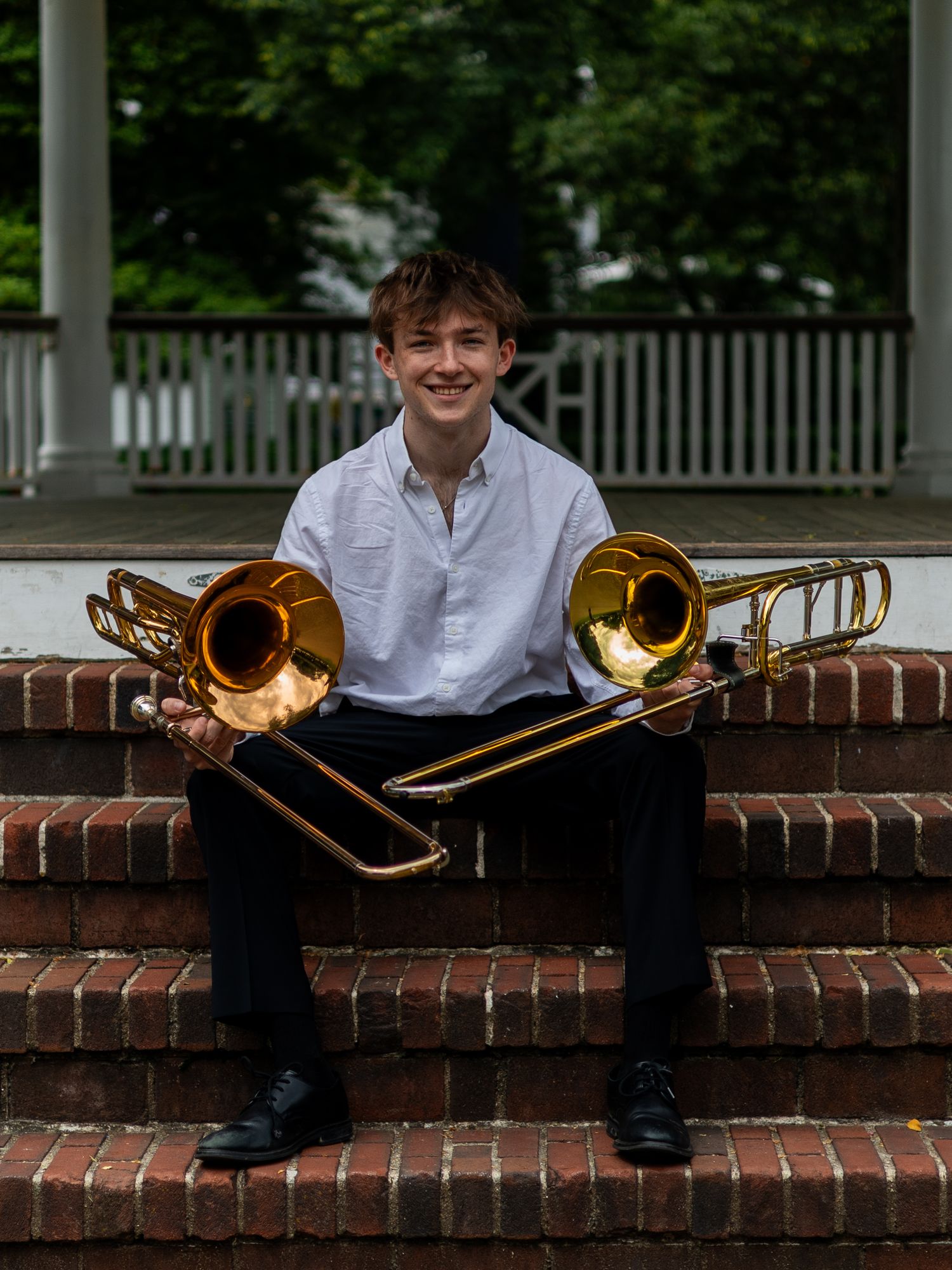 Smiling young man sits on brick steps holding two trombones near a white gazebo outdoors.