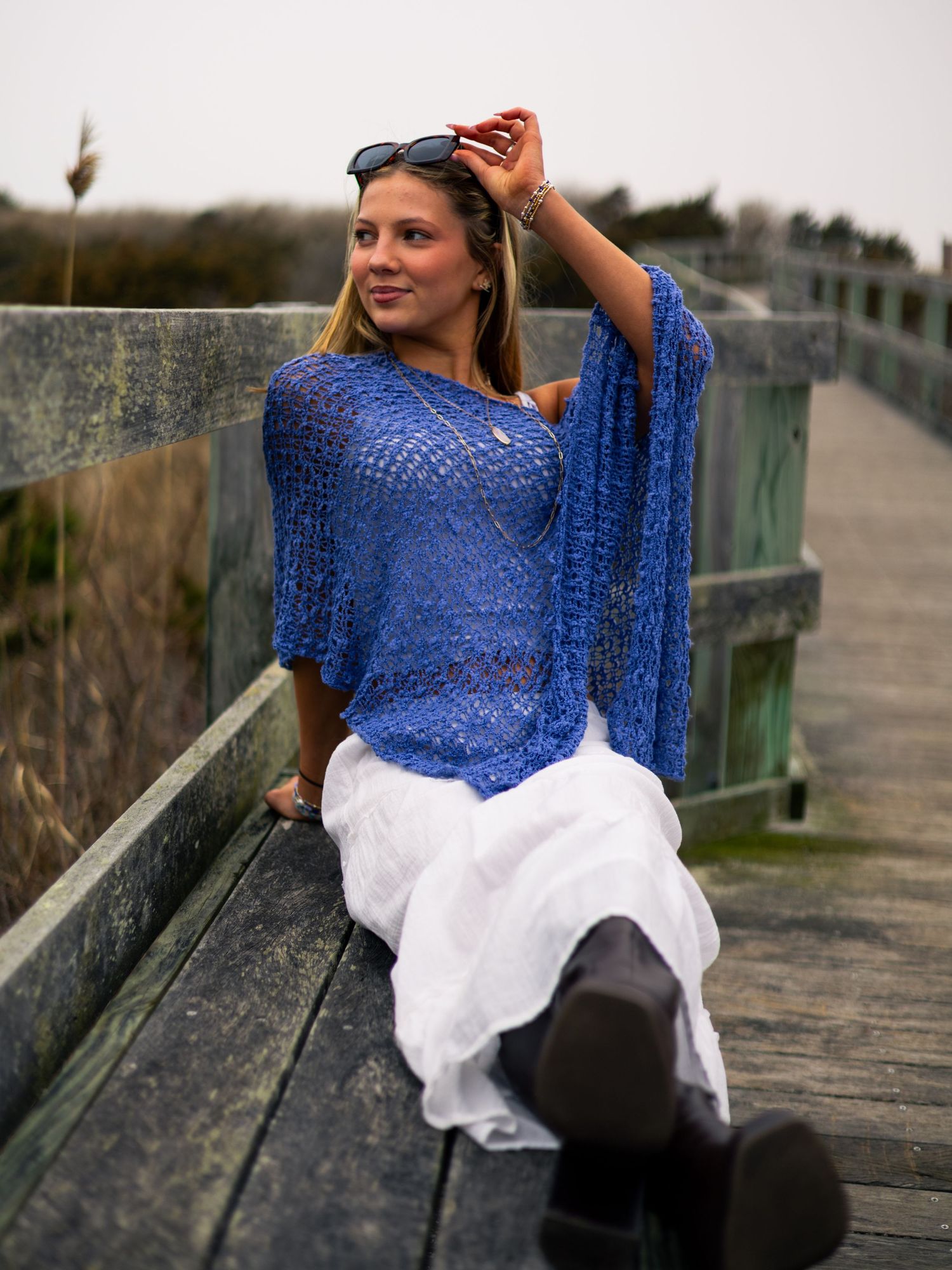 Smiling blonde woman in blue lace top and white skirt sitting on wooden bridge railing outdoors.