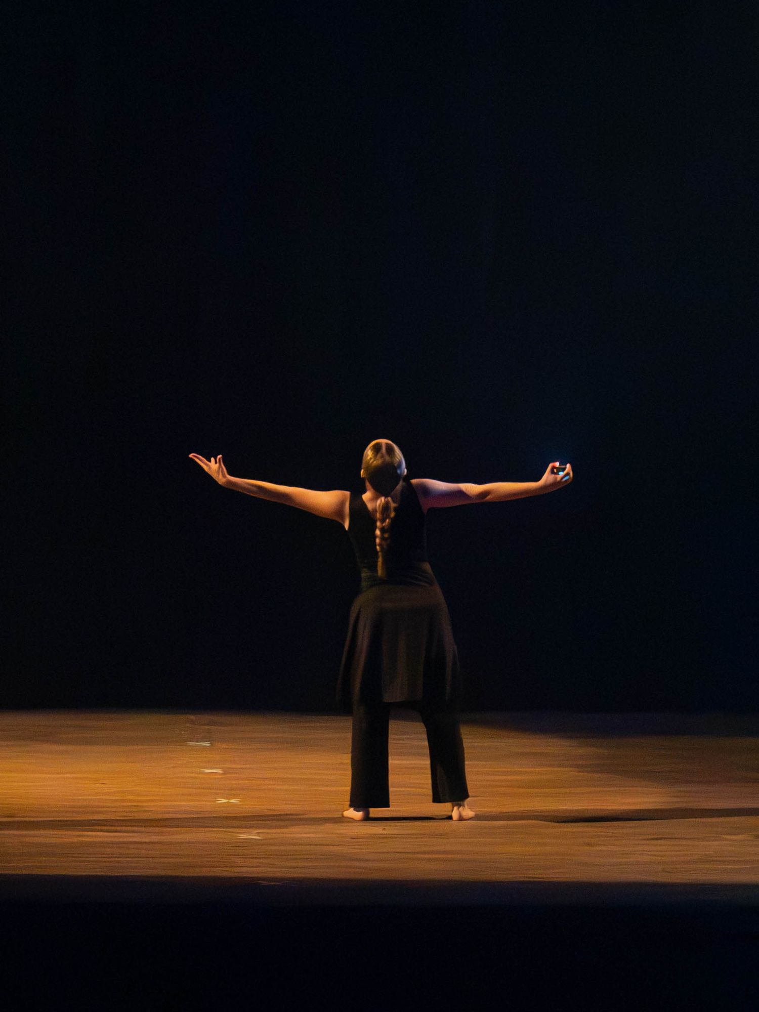 A dancer in black attire stands on a dimly lit stage with arms outstretched, viewed from behind.
