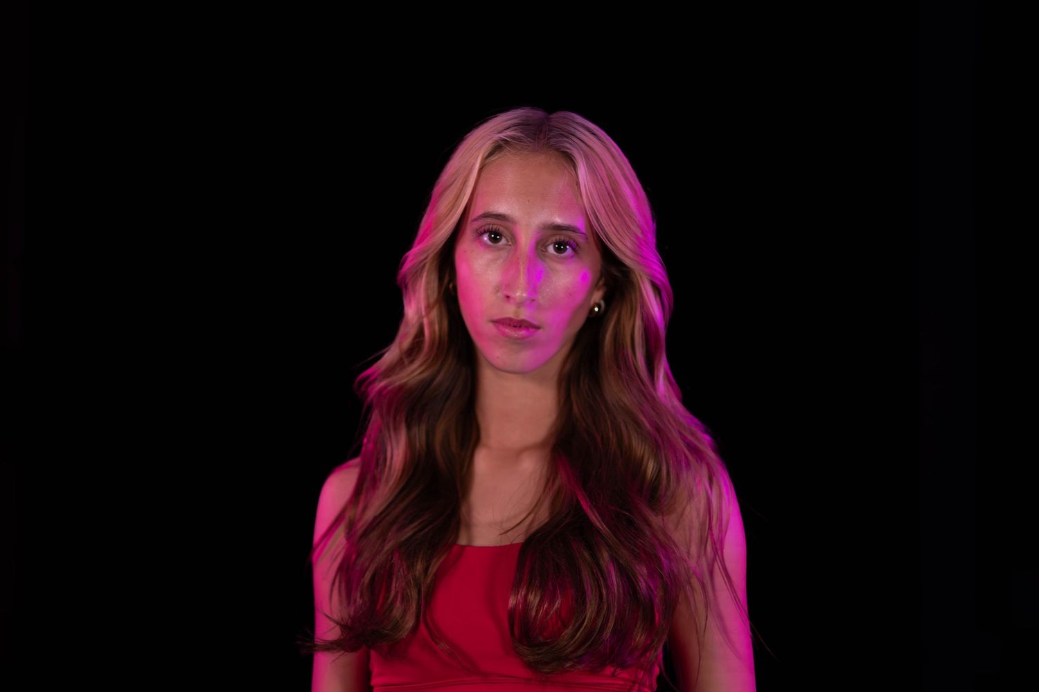 Young woman with long wavy hair and a red top stands against a black background, lit by pink and magenta light.