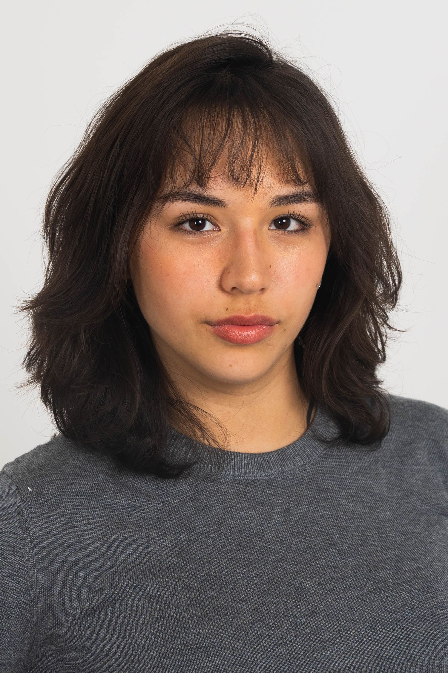 Young woman with shoulder-length dark wavy hair and bangs wearing a gray crewneck sweater against a plain light background.