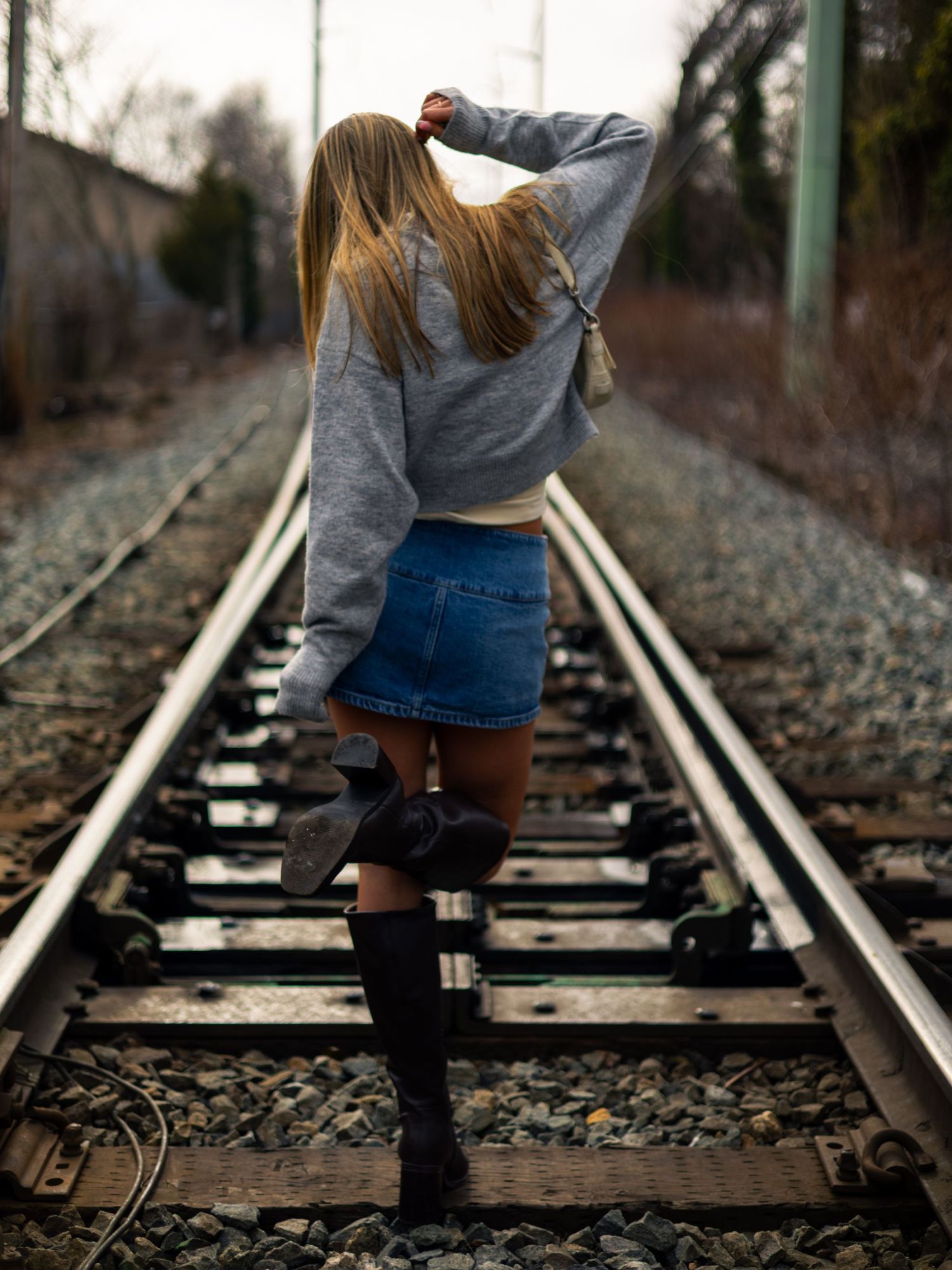 Young woman with long blonde hair walks away along railroad tracks in a gray sweater, denim skirt and black knee-high boots.