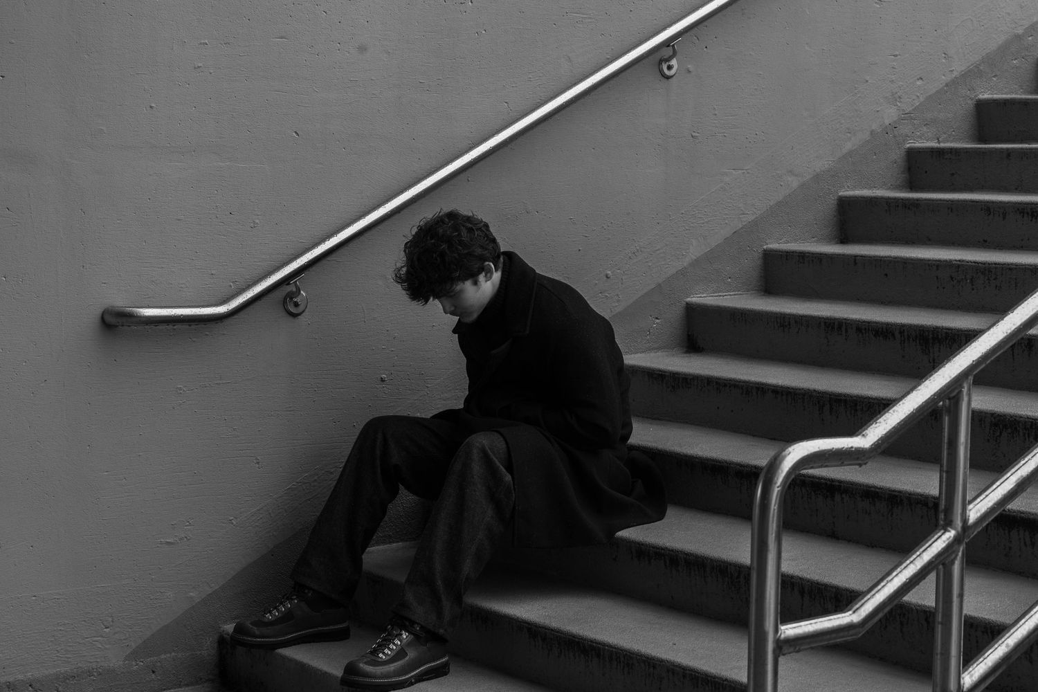 Person in a dark coat sits with head bowed on concrete stairs beside a metal handrail in a moody black and white photograph.