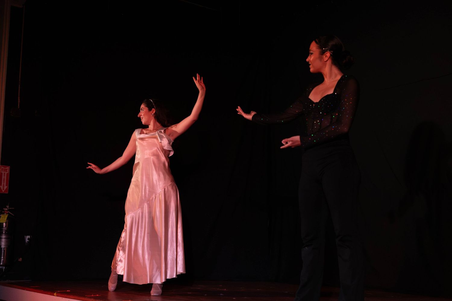 Two women perform on stage, one in a pink gown and one in black, with dramatic gestures under theater lighting.