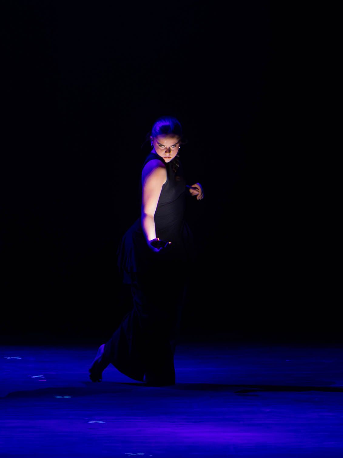 Performer in black dress dancing on a dimly lit stage with dramatic blue and purple spotlight lighting.