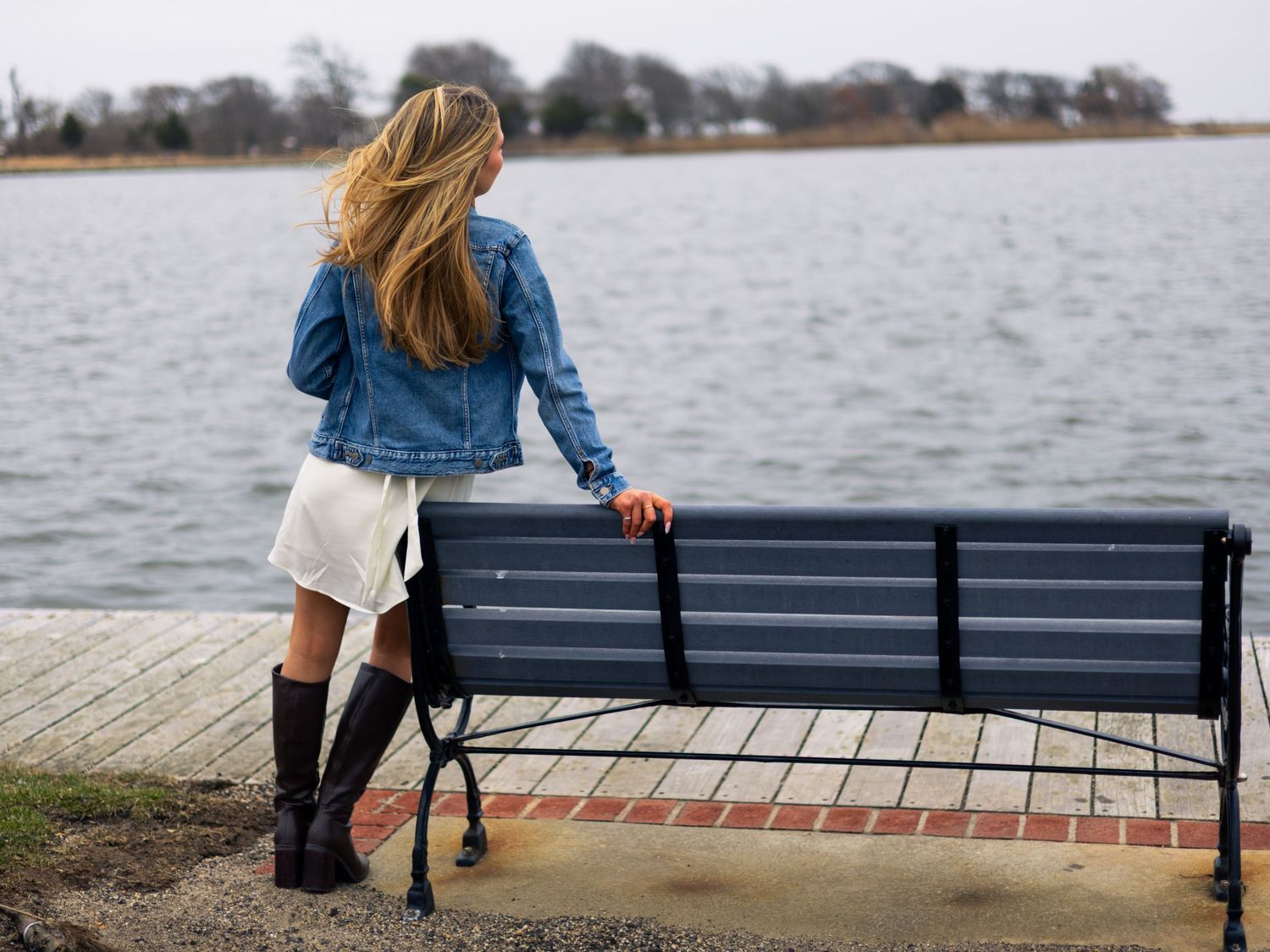 Woman with long blonde hair in a denim jacket, white skirt and black boots stands beside a blue waterfront bench overlooking calm water.
