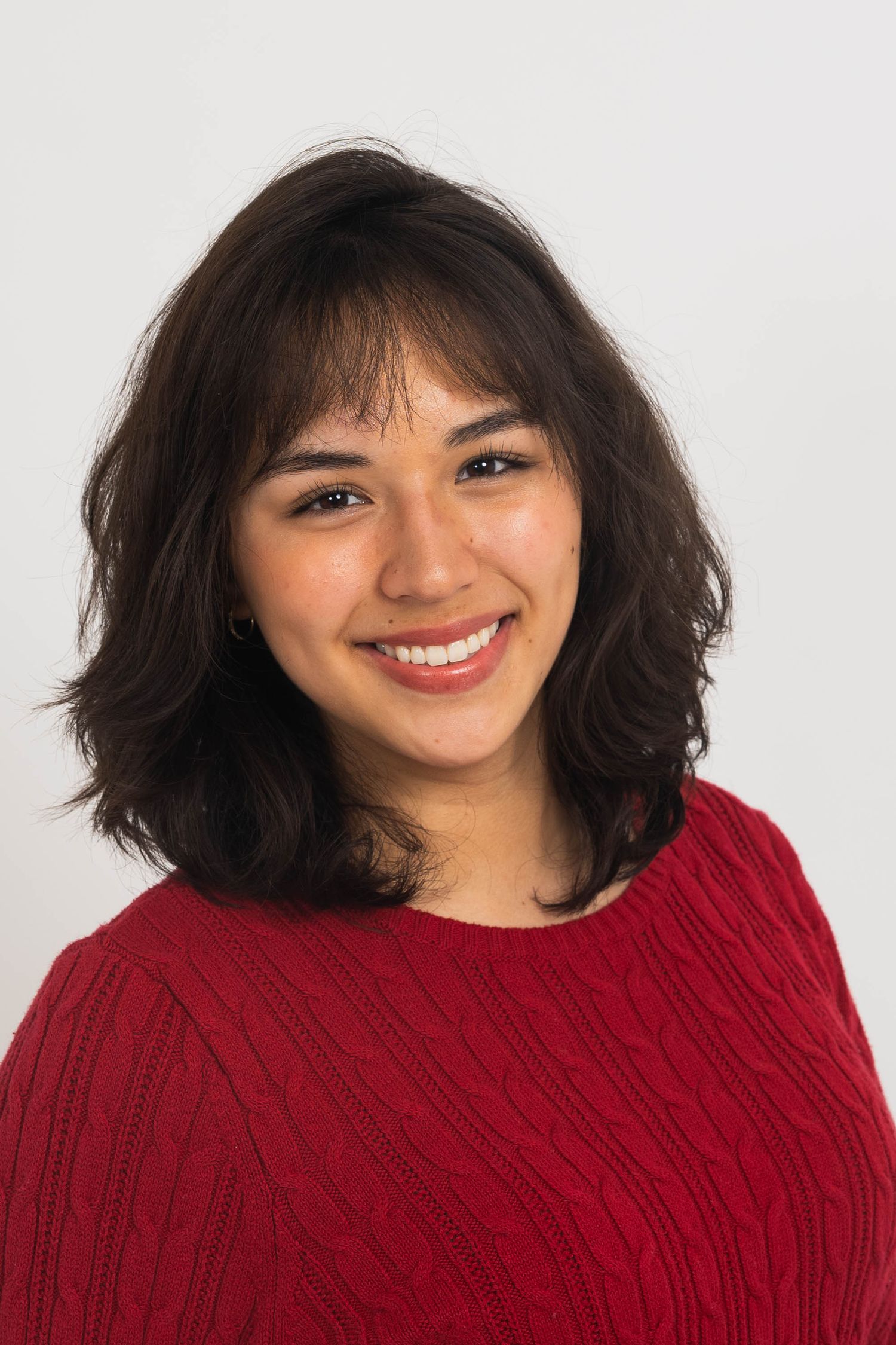 Smiling young woman with shoulder-length wavy dark hair and bangs wearing a red cable-knit sweater against a plain white background.