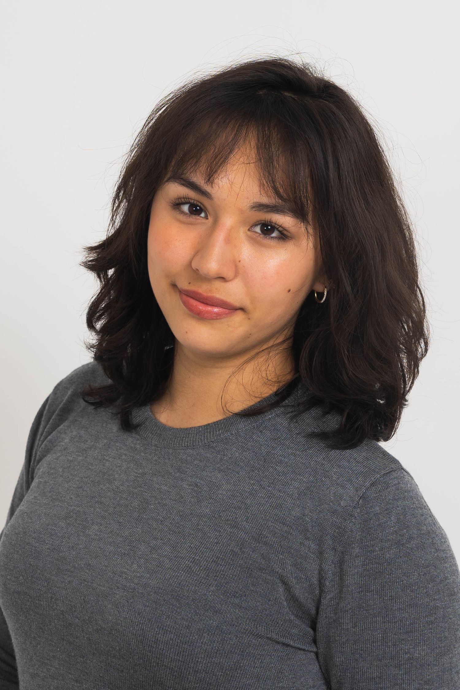 Young woman with shoulder-length dark wavy hair and bangs, wearing a gray sweater and small hoop earring, smiling softly against a plain bac