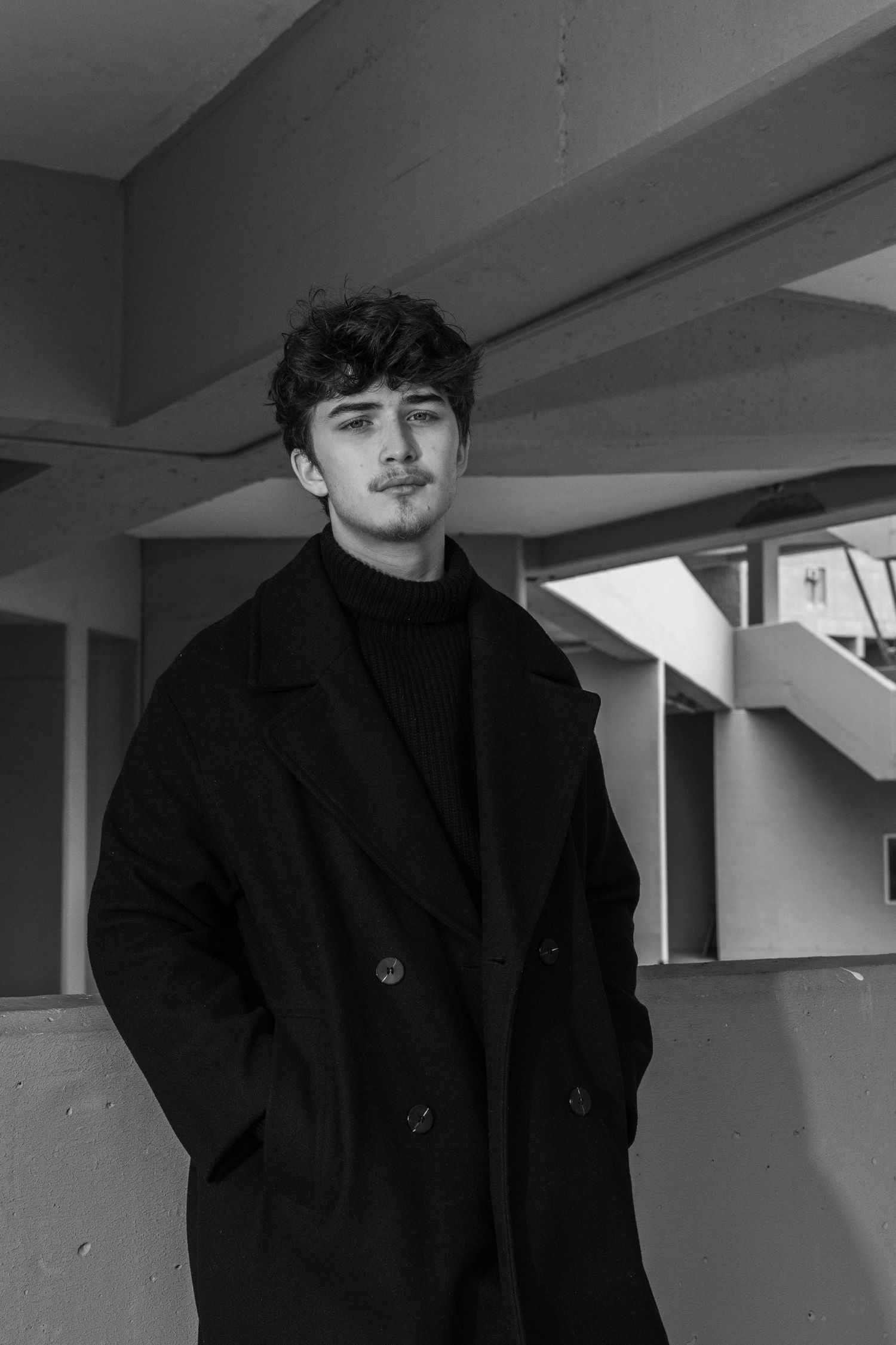 Black and white portrait of a young man with curly hair and a mustache, wearing a dark overcoat and turtleneck in a concrete building.