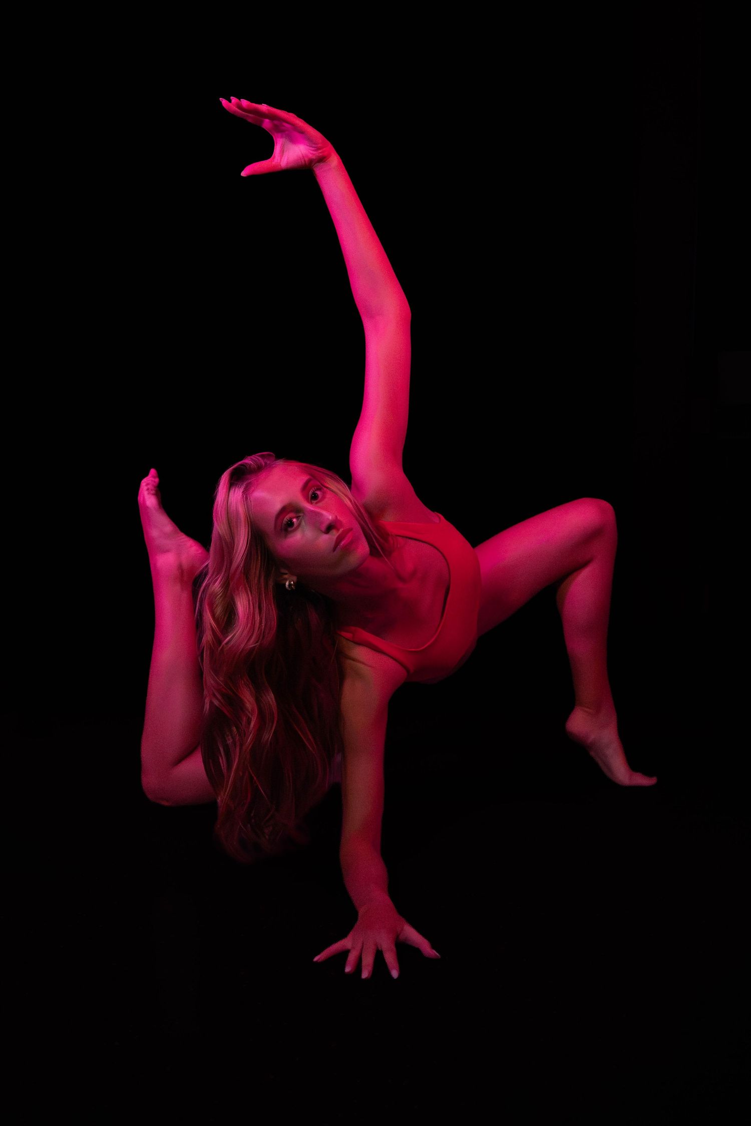 Long-haired dancer in a red leotard balancing on one hand with an arm stretched high, bathed in crimson light against black.