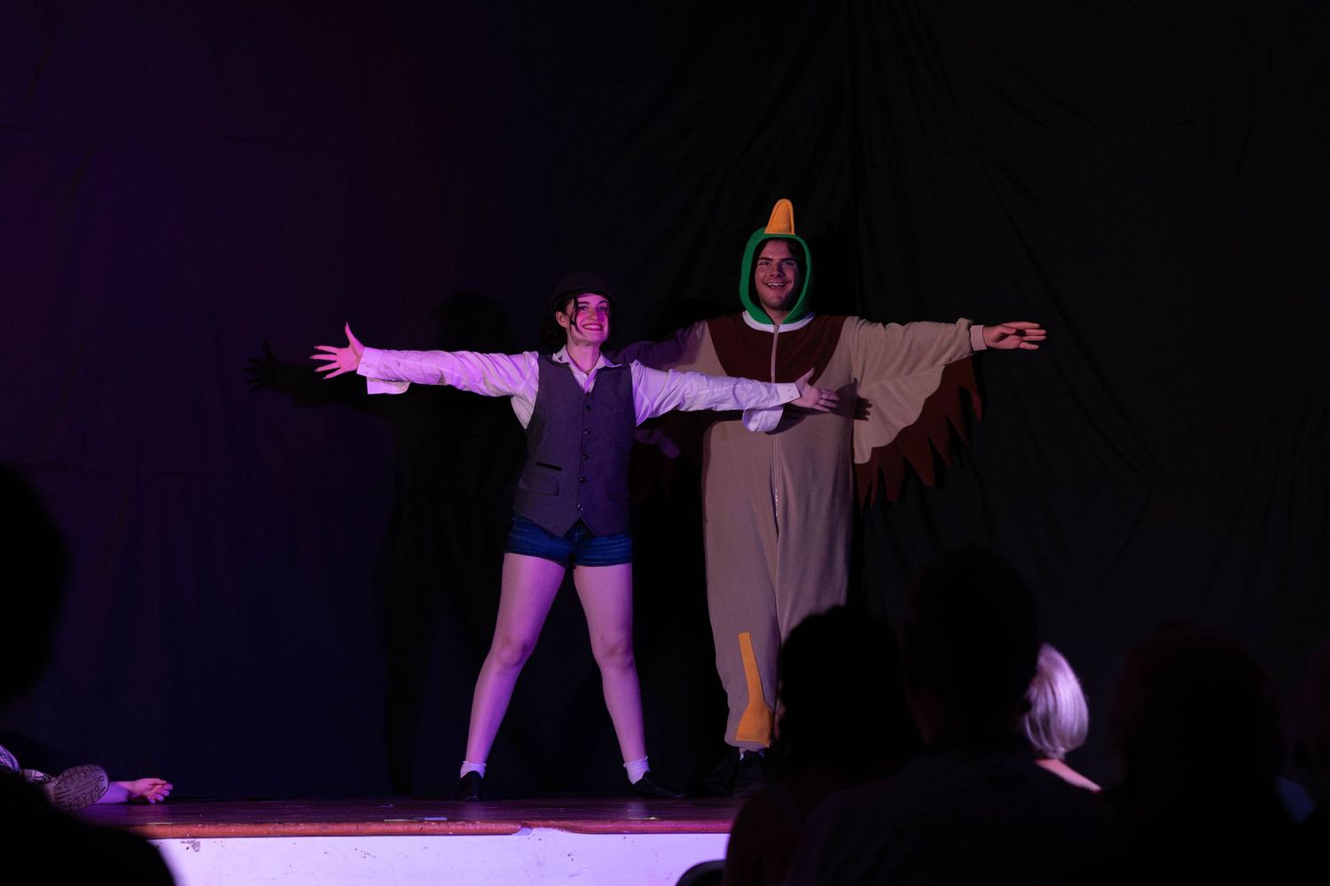 Two costumed performers bow with arms outstretched on a darkened theater stage.