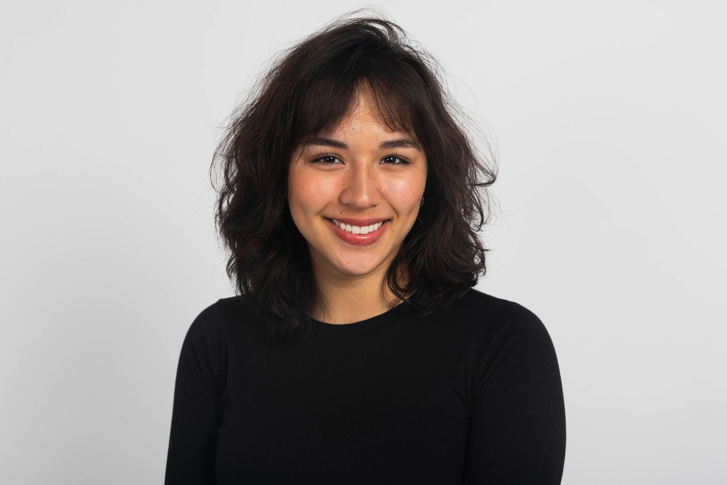 Smiling young woman with dark wavy shoulder-length hair and bangs wearing a black long-sleeve top against a plain light gray background.