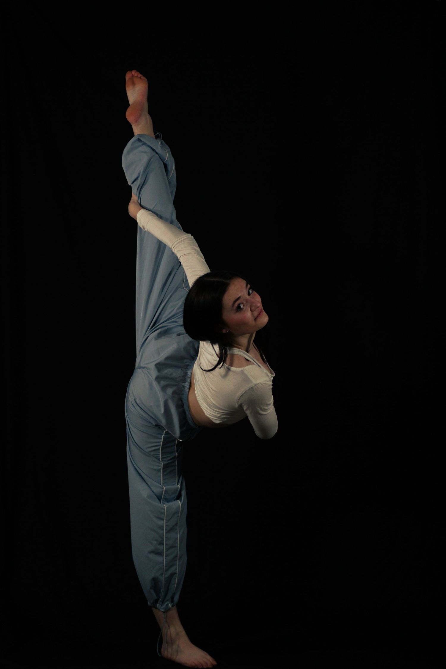 Young dancer in a light blue pants and white top performs a vertical standing split against a black studio background.