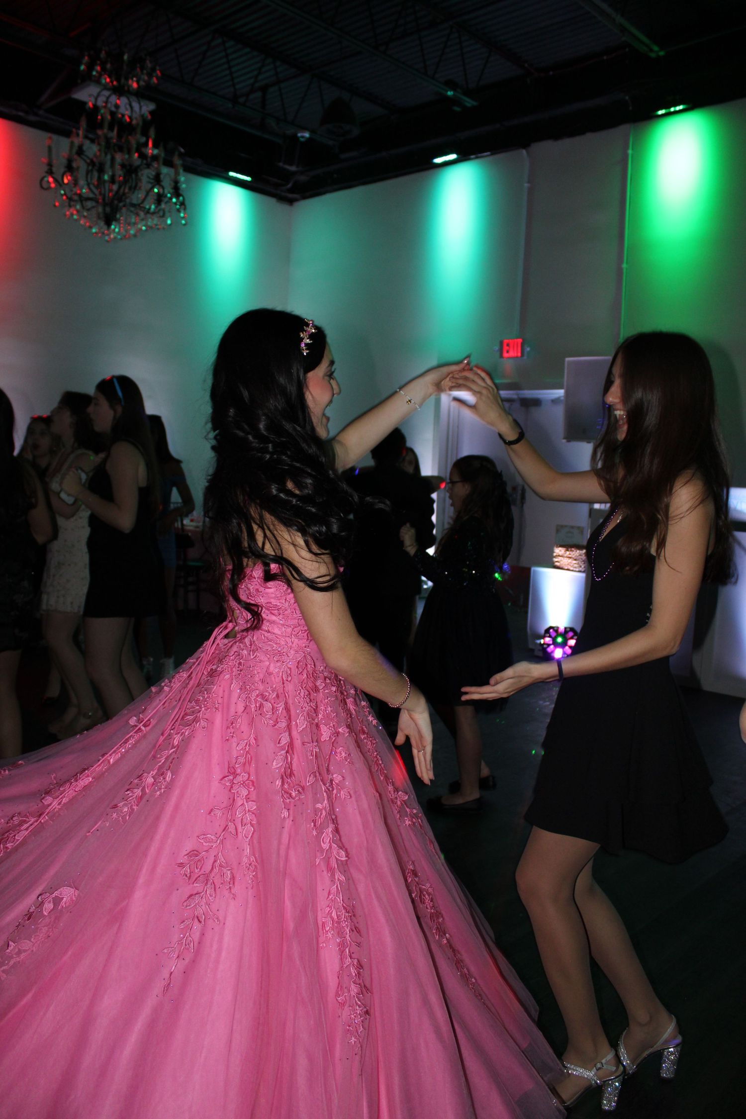 Girl in pink ball gown dancing with friend in black dress at a quinceañera party.