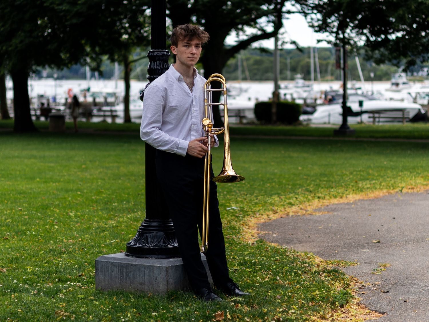 Young trombone player posing beside park pedestal with marina visible in background.