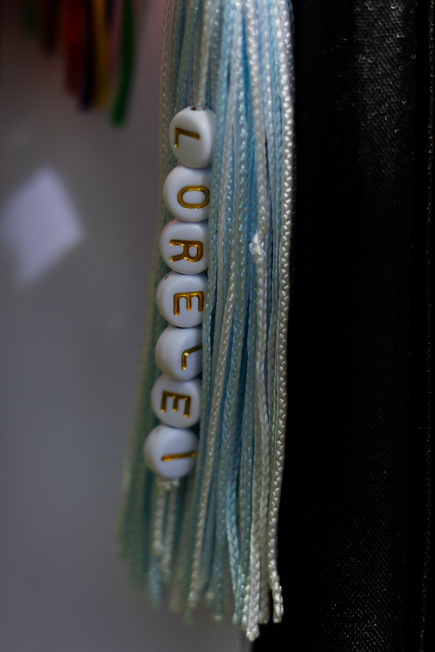 Graduation cap tassel with white letter beads spelling LORELEI on a light blue decorative cord.