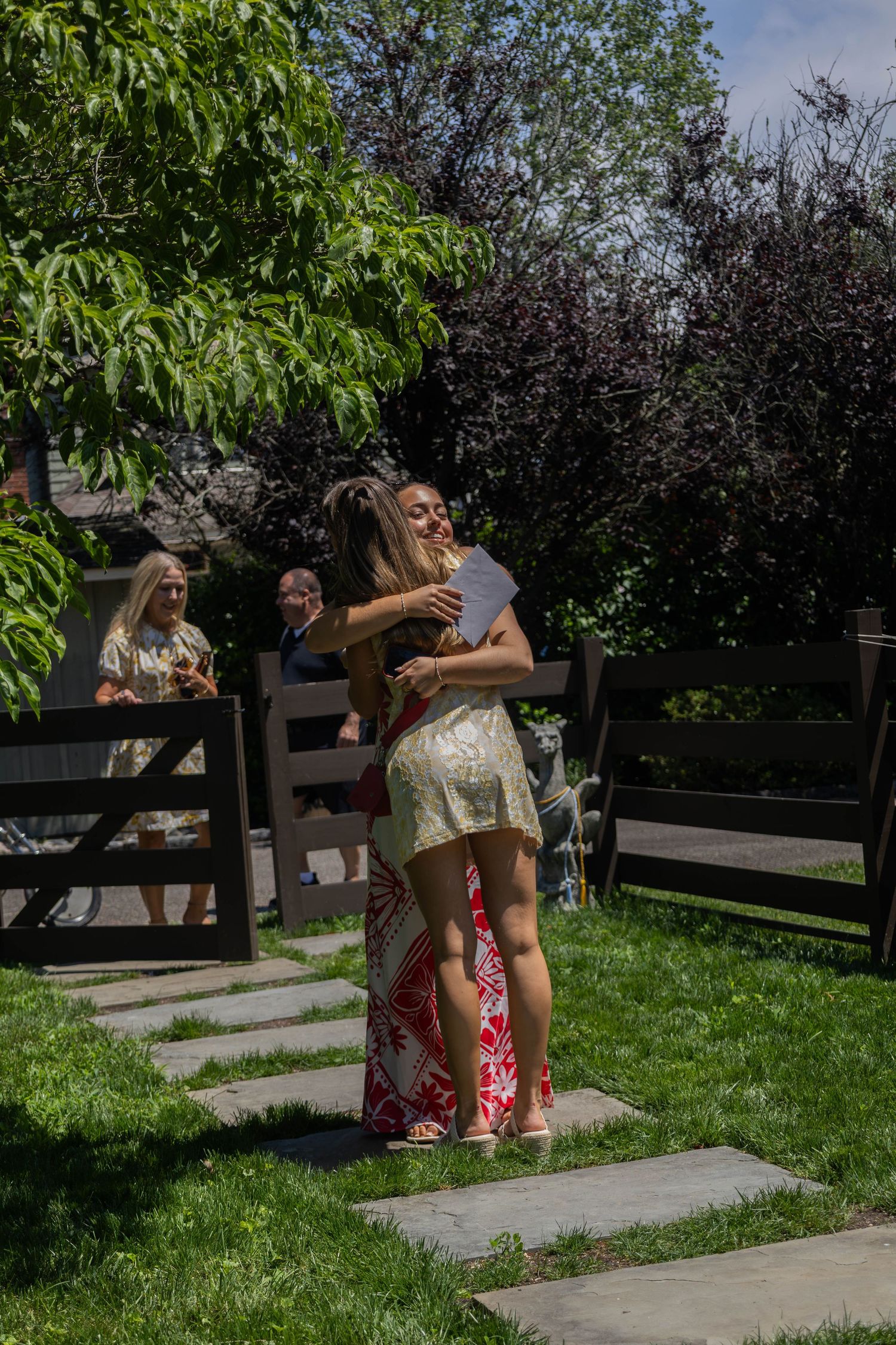 Two young women share a warm embrace outdoors in a sunny garden during a graduation celebration party.