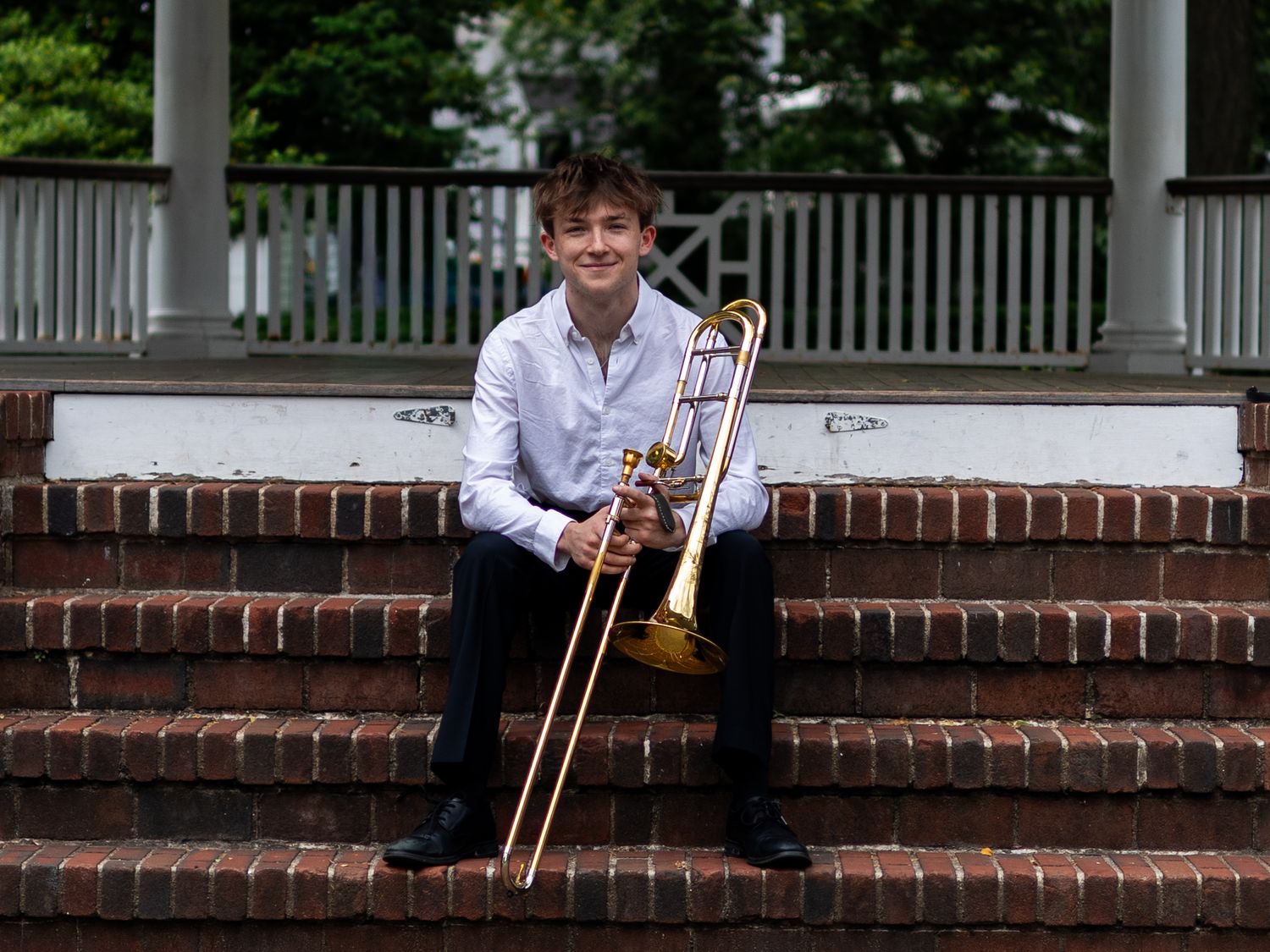 Teenage trombone player sitting on brick steps outside white gazebo, smiling at camera.