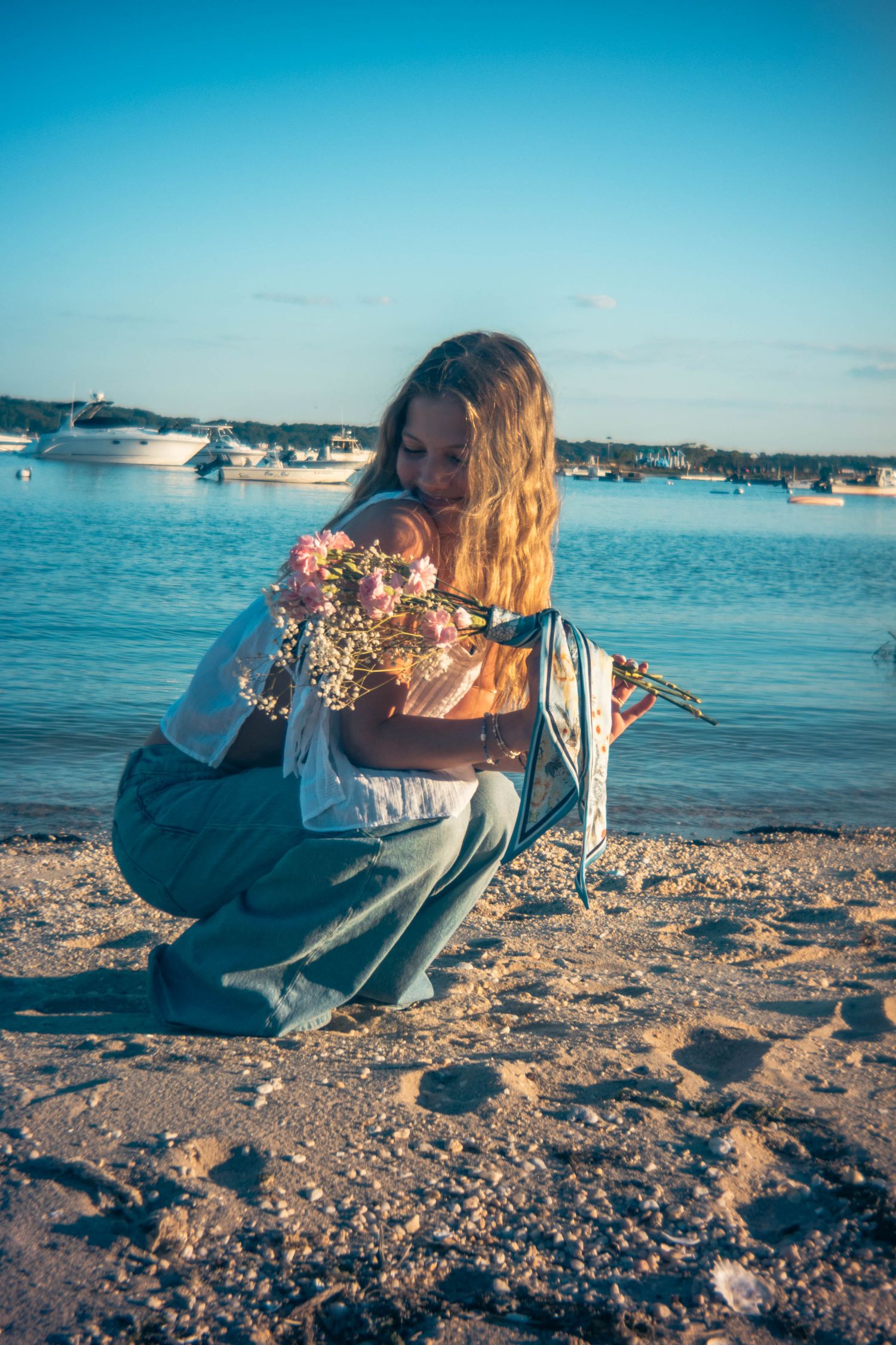 Woman with long hair crouching on a sandy beach holding a pink flower bouquet, with boats on calm blue water behind her.
