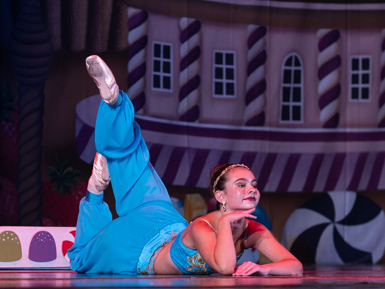 Ballerina in blue costume performs elegant floor pose on stage with colorful candy-themed backdrop during ballet performance.