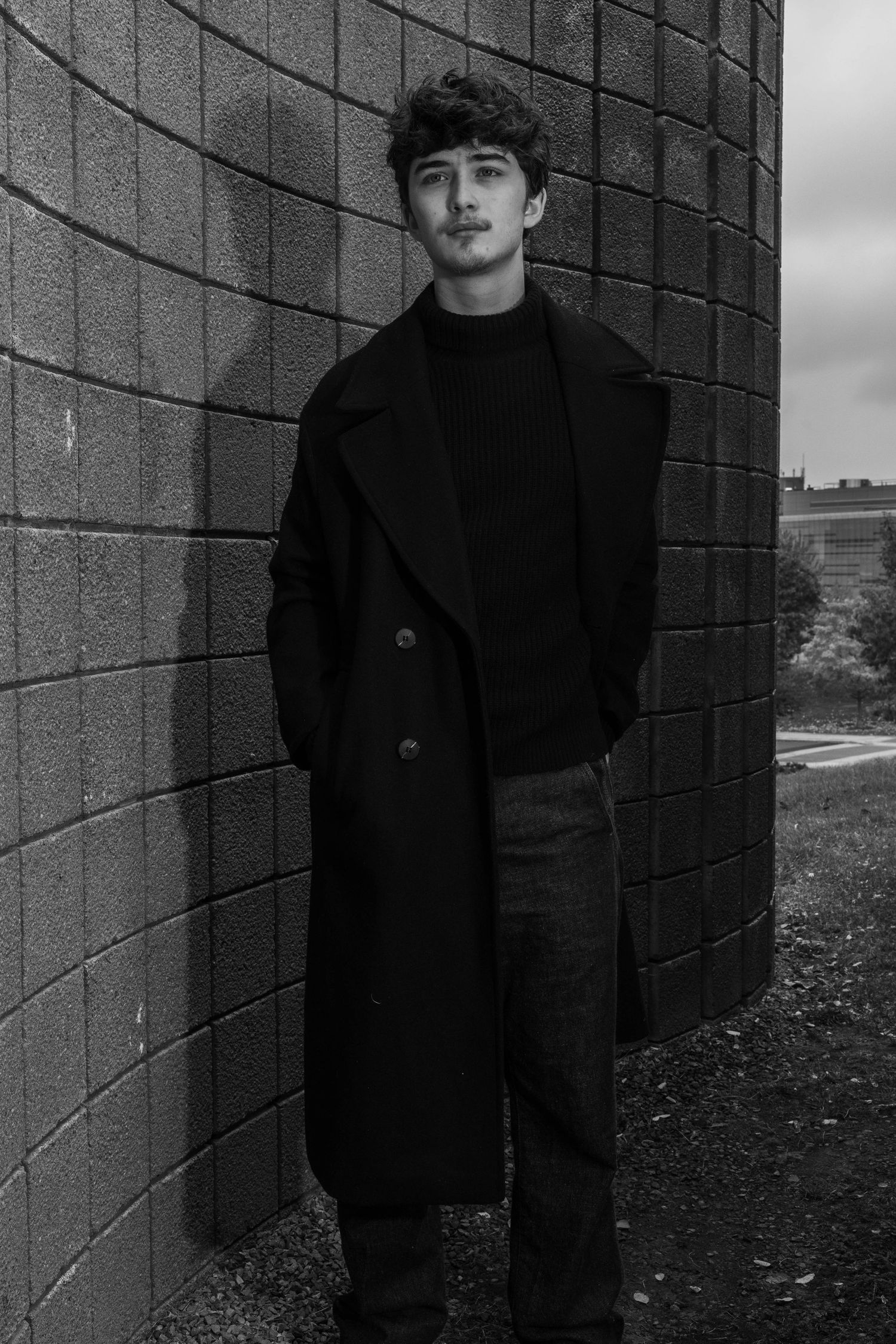 Black and white portrait of a young man with dark curly hair in a long coat and turtleneck, standing against a curved brick wall.