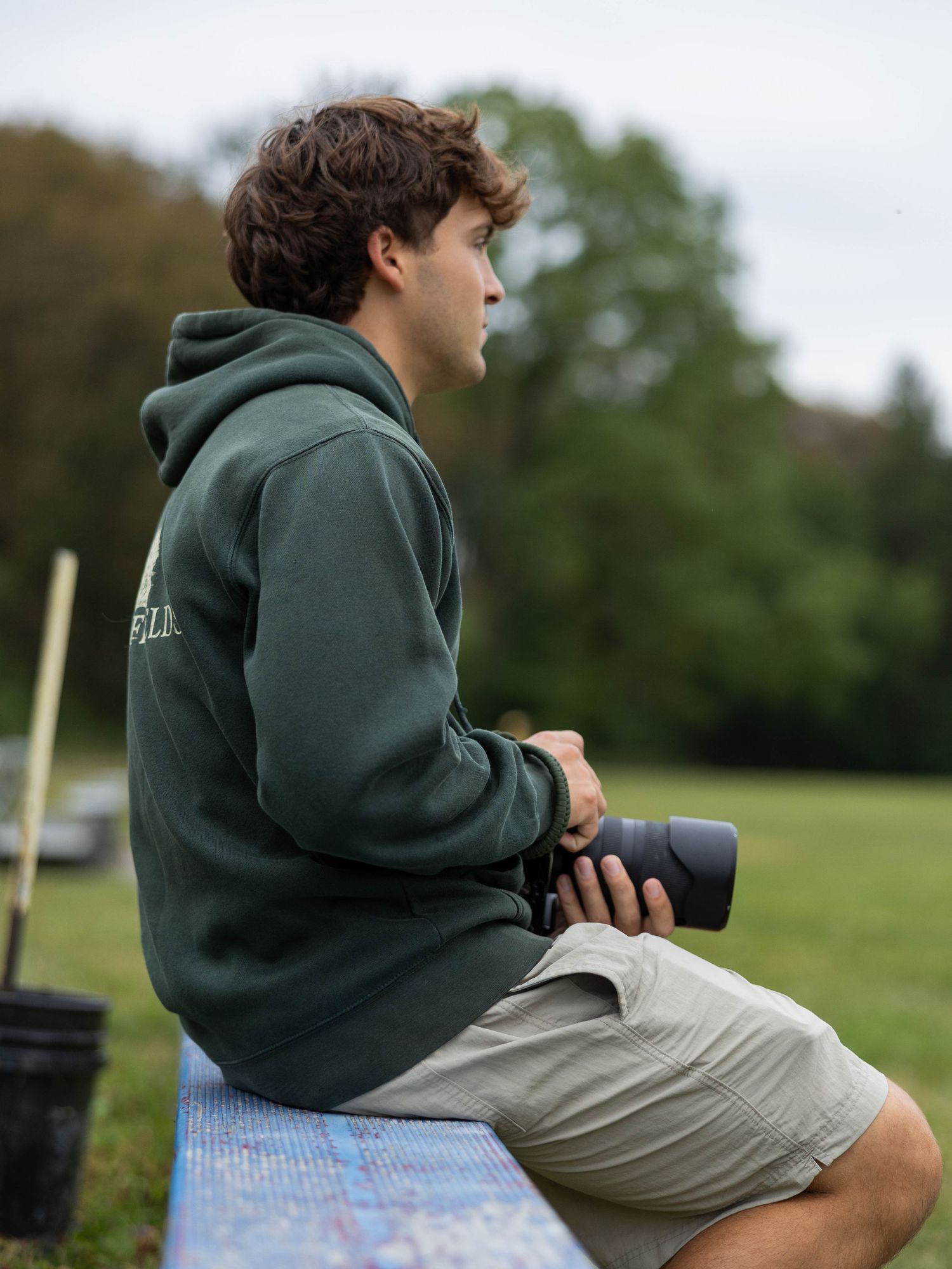 Young man in green hoodie and khaki shorts sits outdoors holding a camera, gazing into the distance.