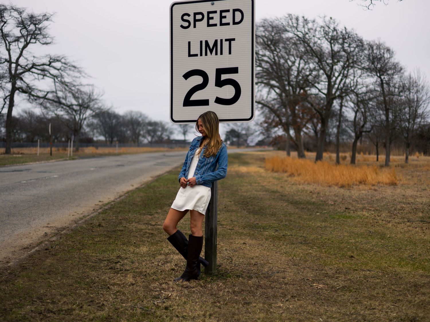 Young woman in denim jacket and white skirt leaning against a speed limit 25 sign on a rural road.