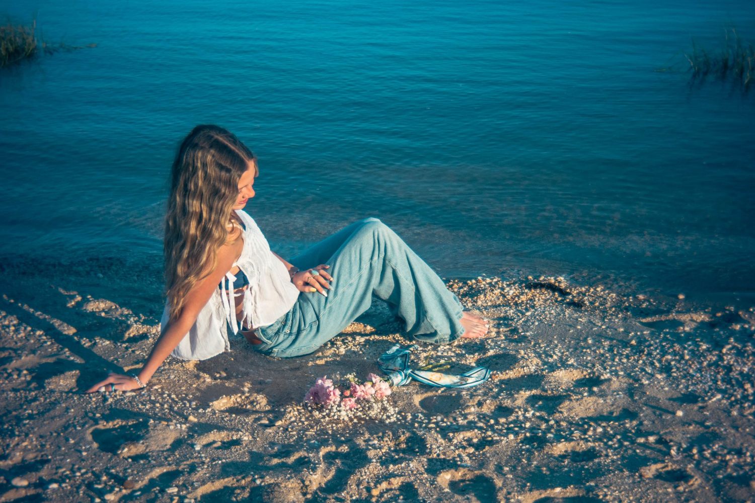 Woman in white top and teal wide-leg pants relaxing on pebble beach beside calm blue water.