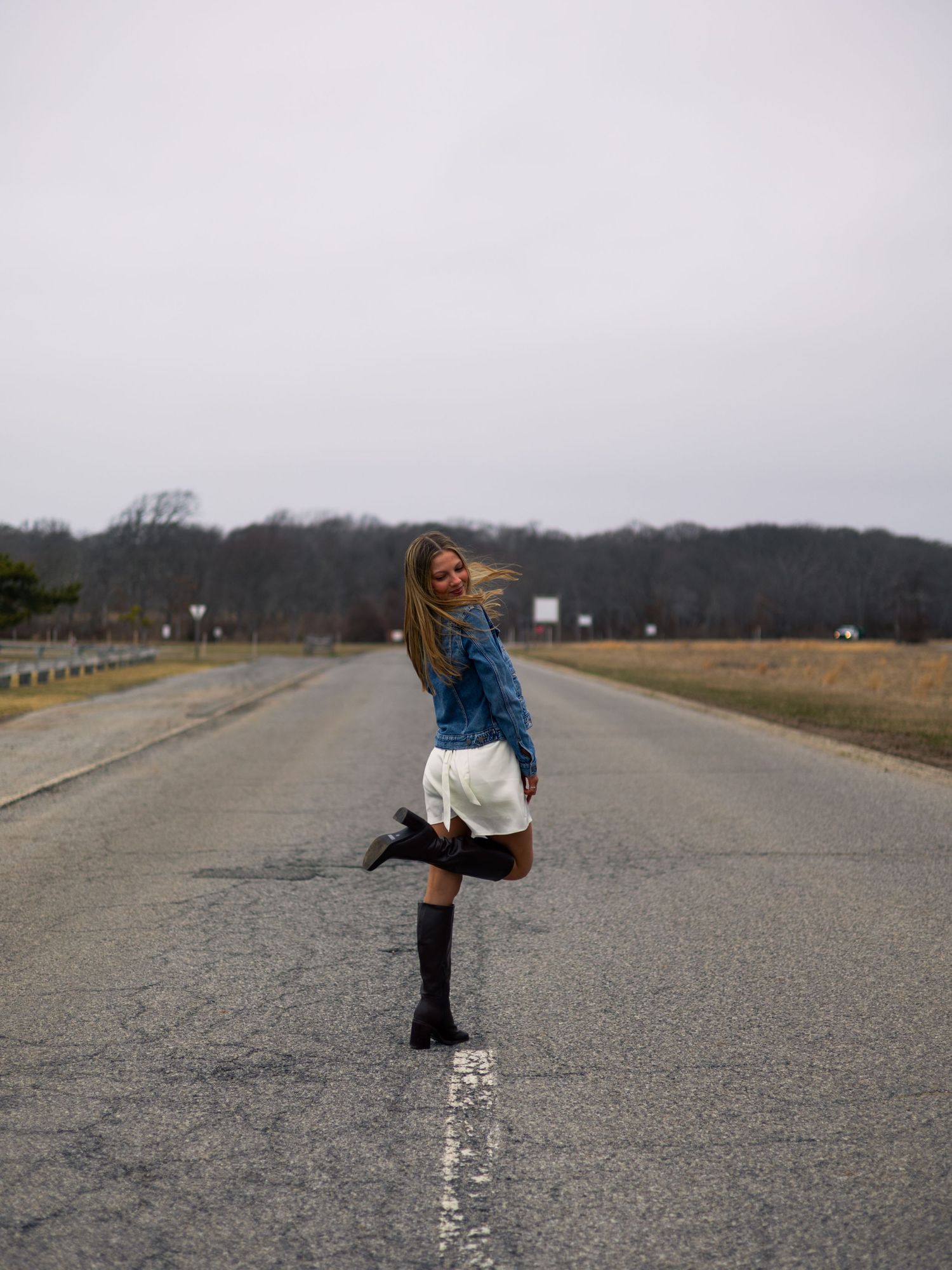 Young woman in white skirt and denim jacket twirling on empty road with knee-high black boots.