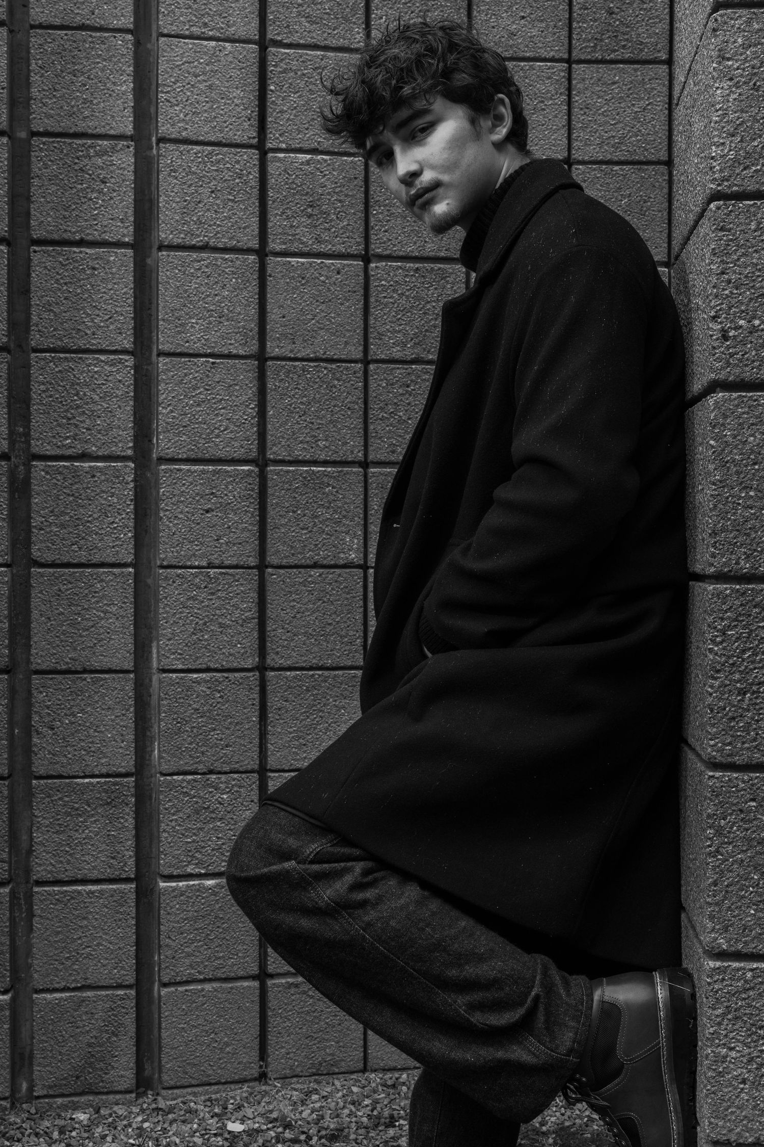 Black and white portrait of a curly-haired young man in a long dark coat leaning against a concrete block wall with one boot raised.