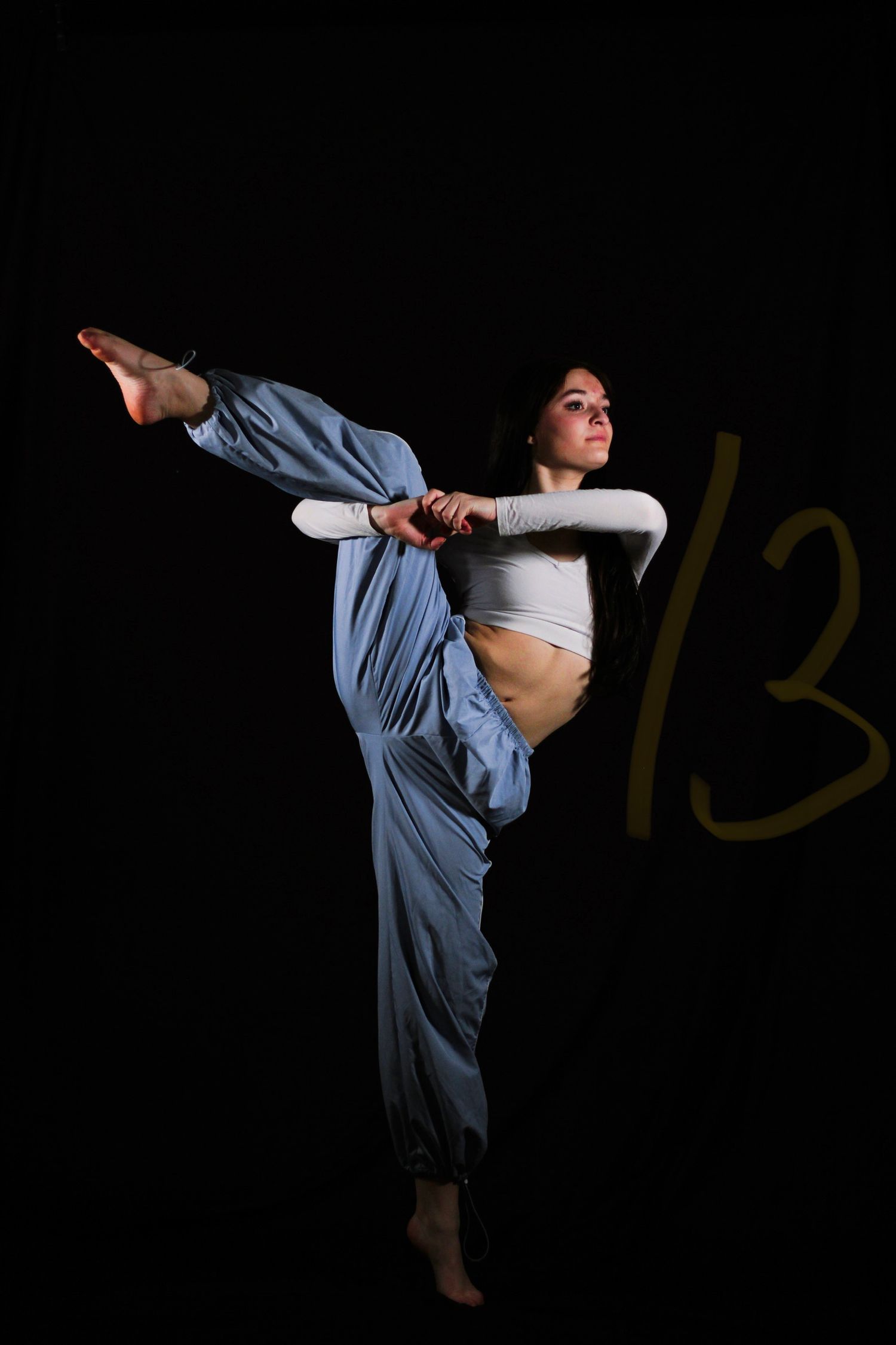 Barefoot dancer in baggy blue pants and a white crop top performs a standing split with one leg raised high against a black backdrop.