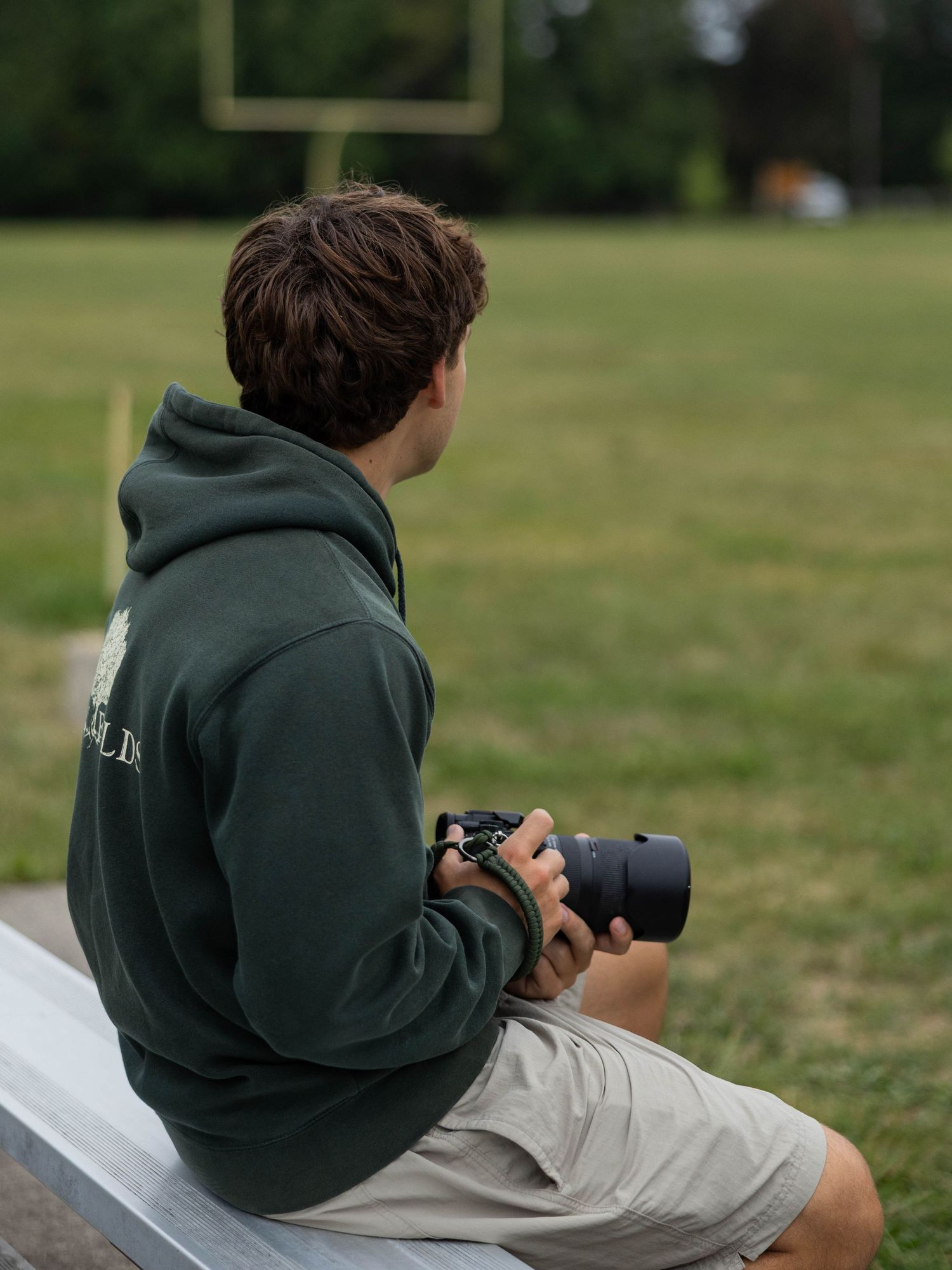 Young man in green hoodie sitting on bleachers holding a camera, overlooking a football field.