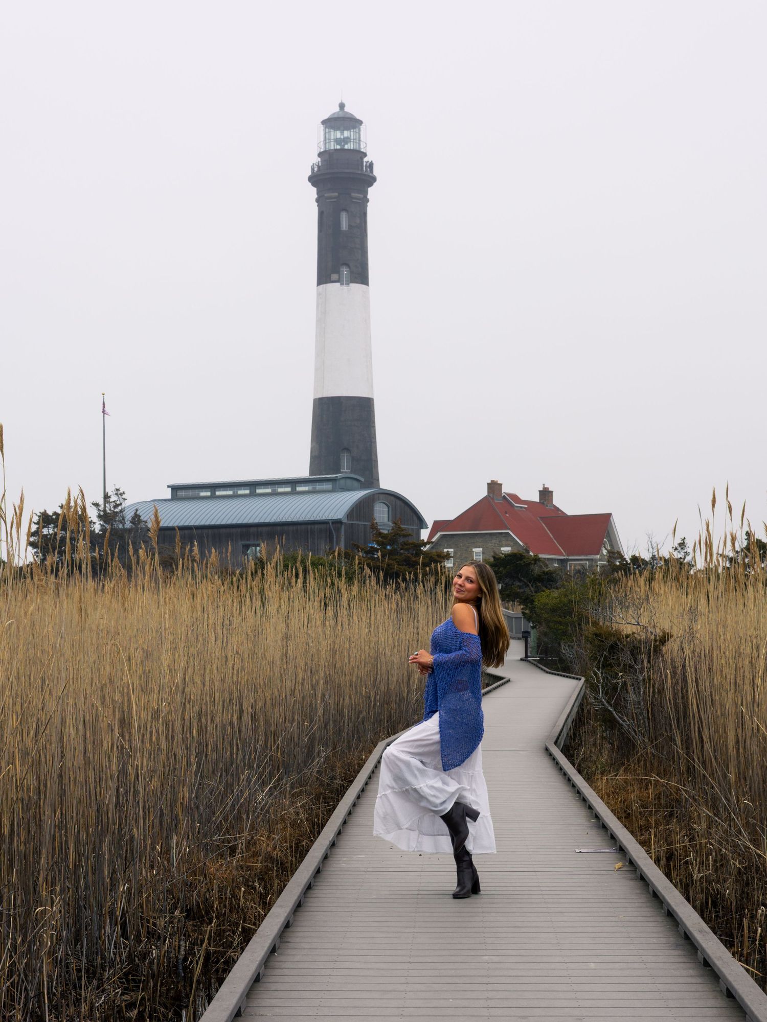 Woman in blue top and white skirt poses on boardwalk path leading to Fire Island Lighthouse on overcast day.