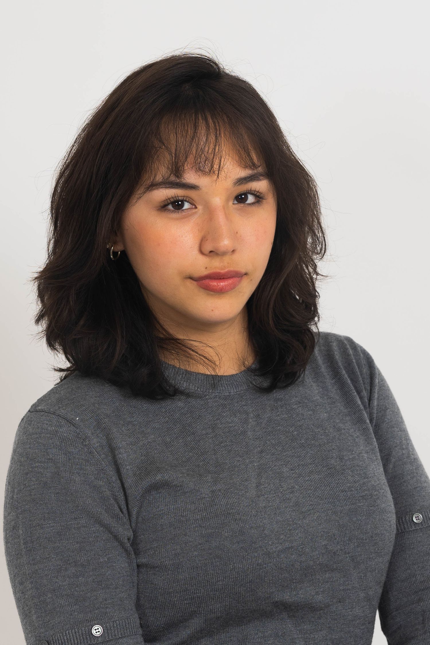 Portrait of a young woman with wavy dark hair, wispy bangs and a small gold earring, wearing a gray sweater, gazing at the camera.