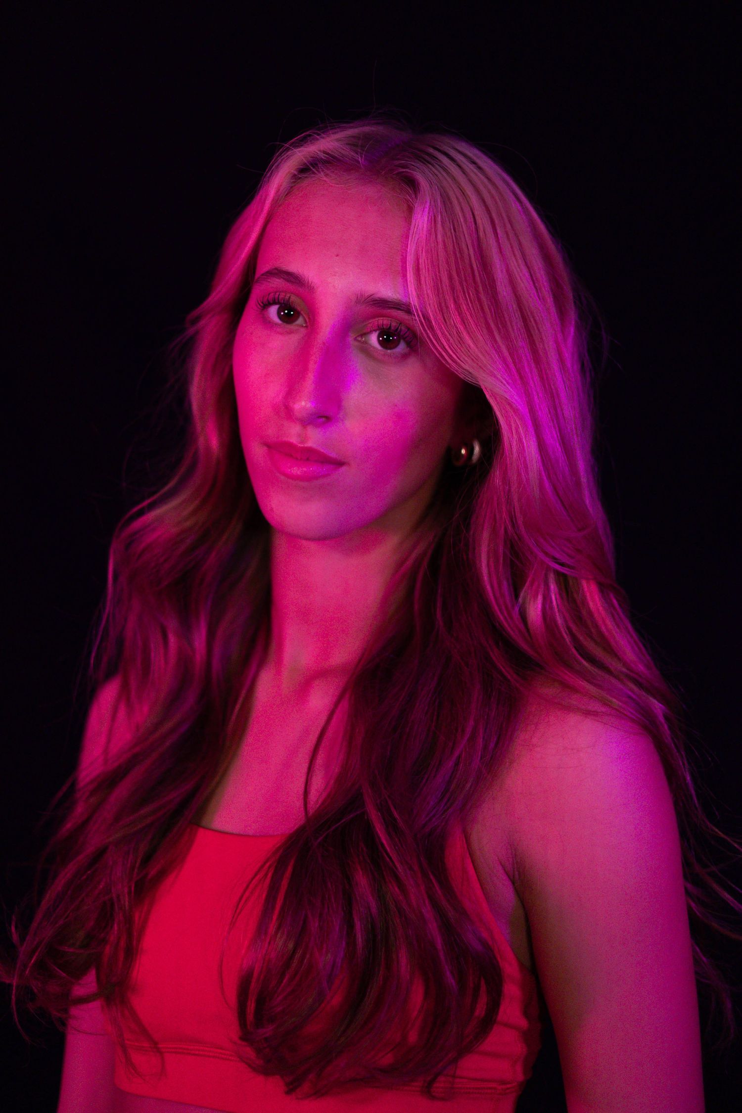 Portrait of a young woman with long wavy blonde hair lit in vivid pink and magenta light against a dark background.