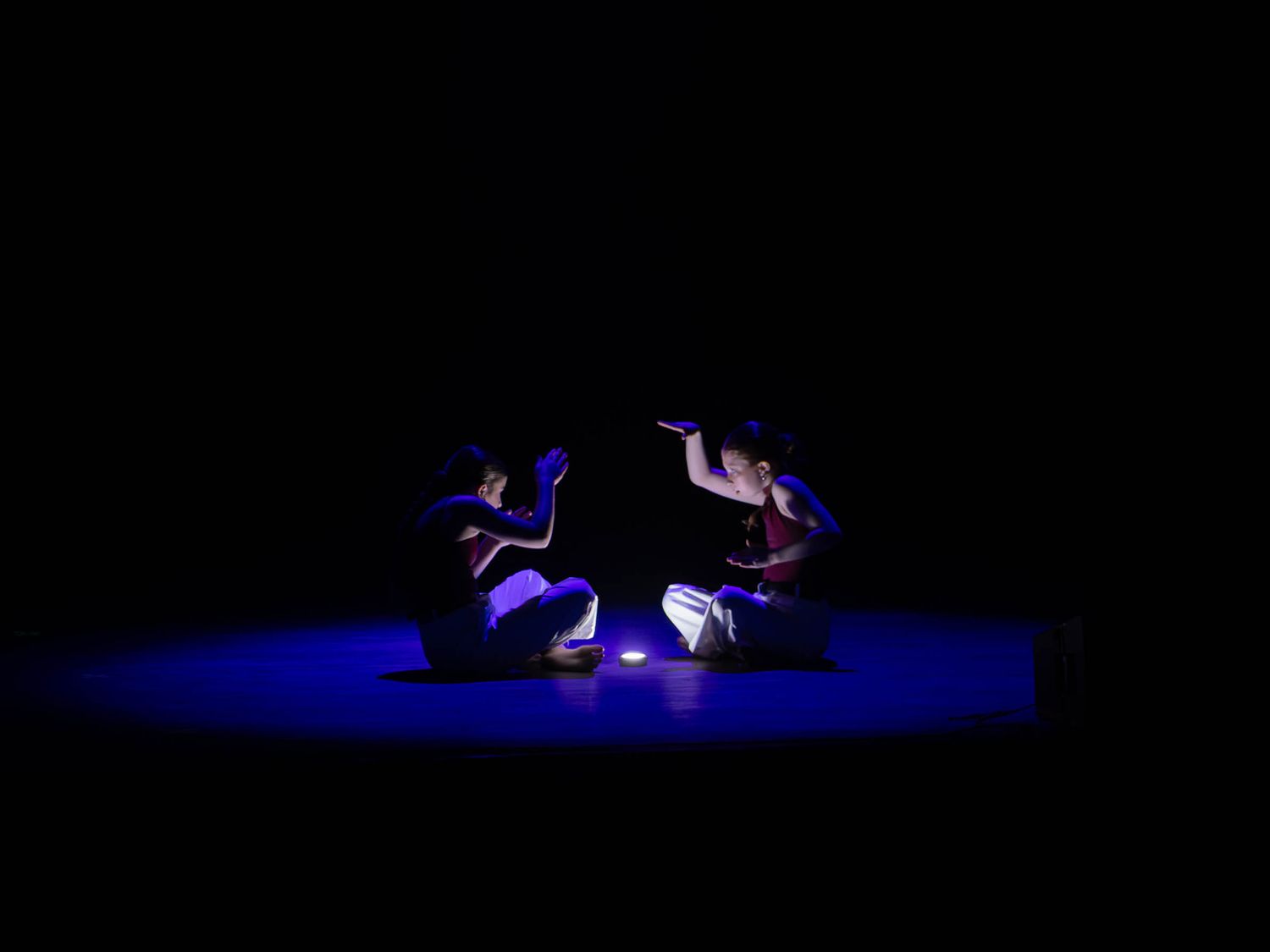 Two dancers sit facing each other in blue stage lighting with a small light between them.