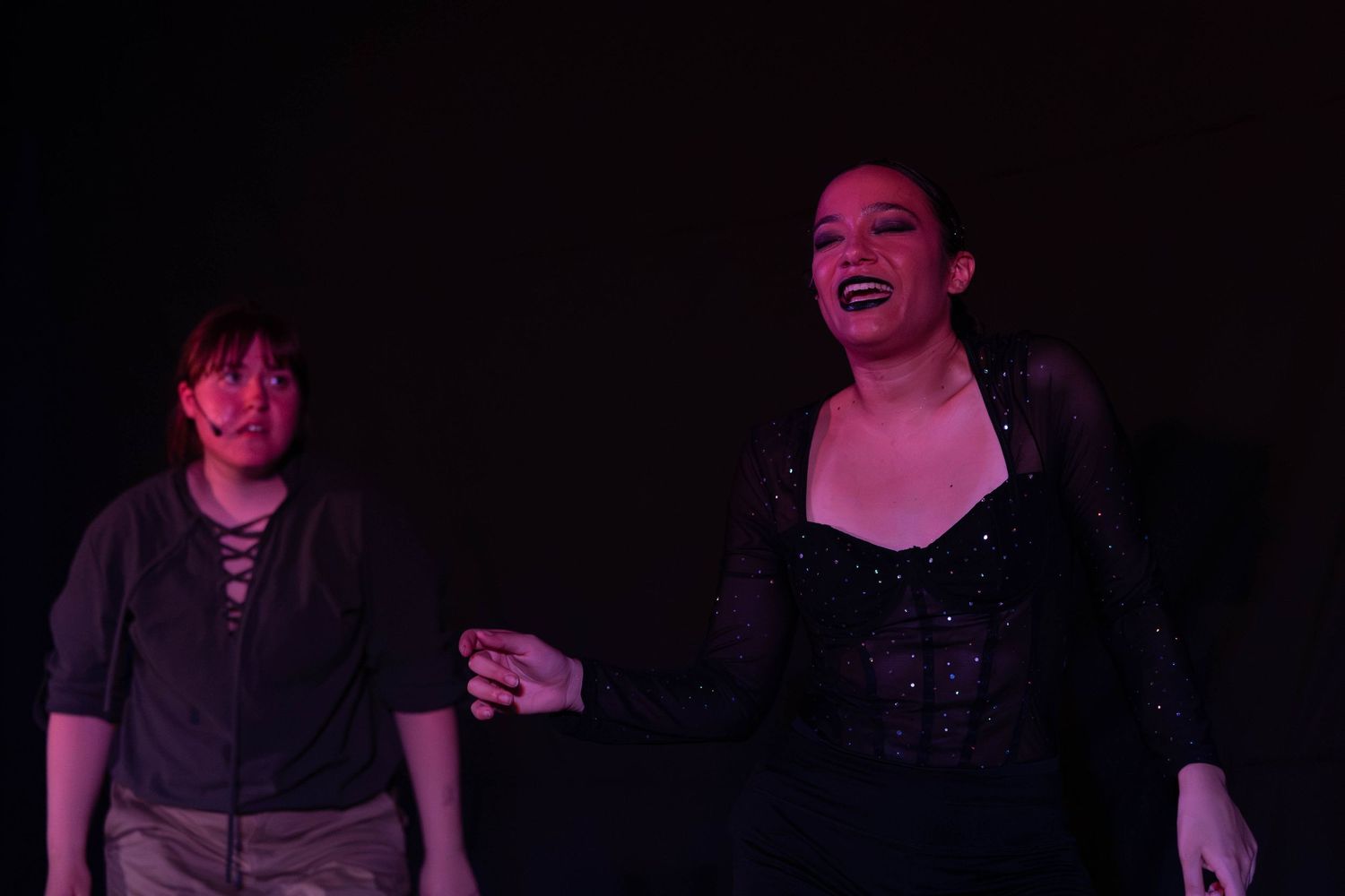 Two performers on a dark stage, one in a black sequined top singing expressively under purple stage lighting.