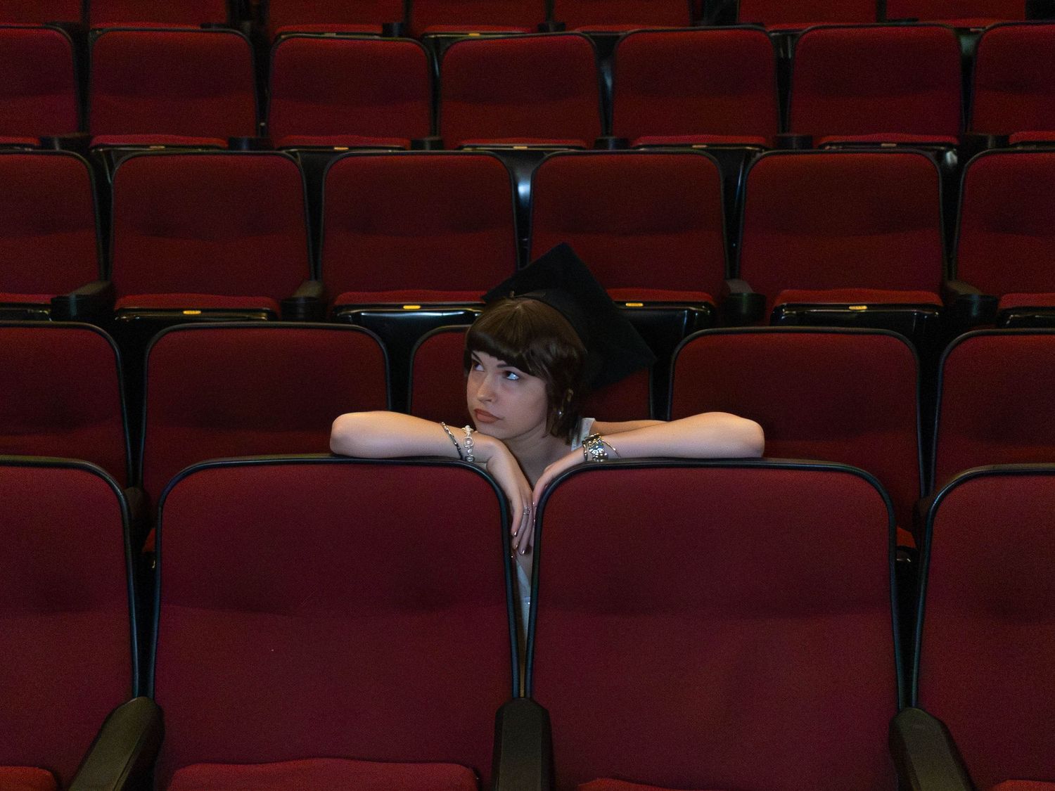 Young graduate in black cap and gown sits alone among rows of empty red auditorium seats.