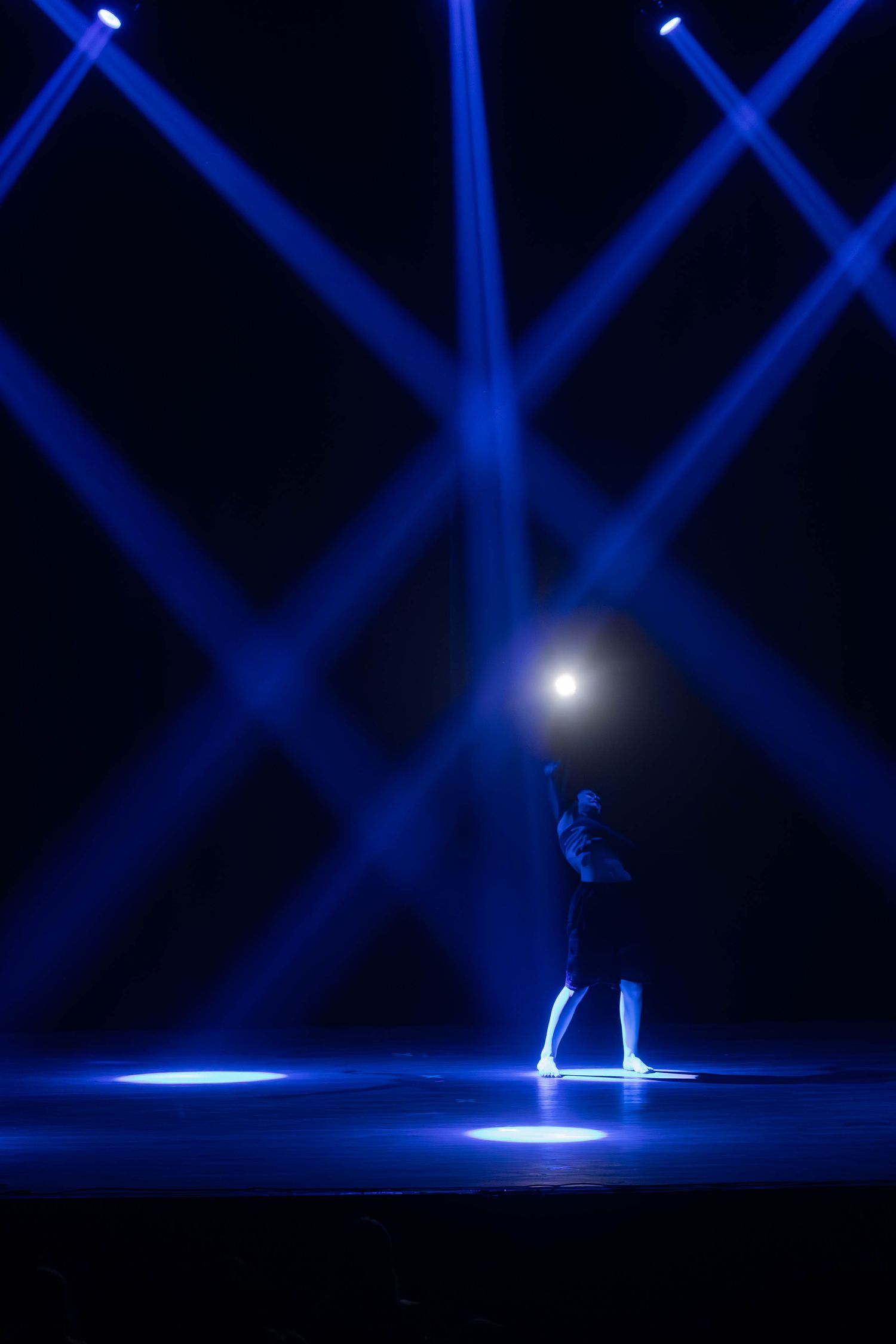 A lone performer stands on a dark stage illuminated by dramatic blue laser beams crossing overhead.